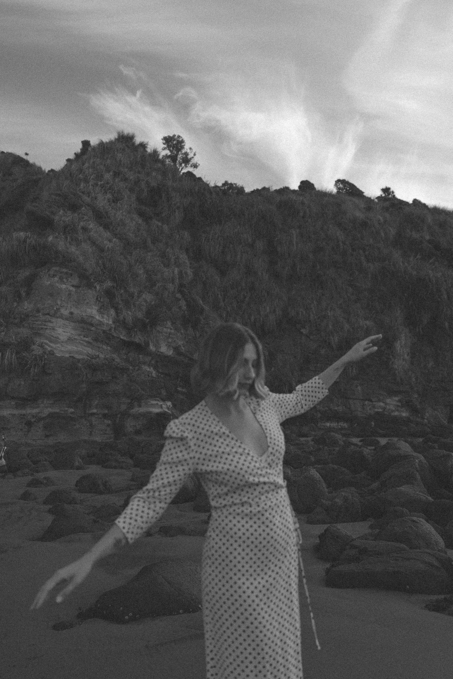 Black and white photo of a woman in a polka dot dress posing with outstretched arms on a rocky beach in New Zealand.