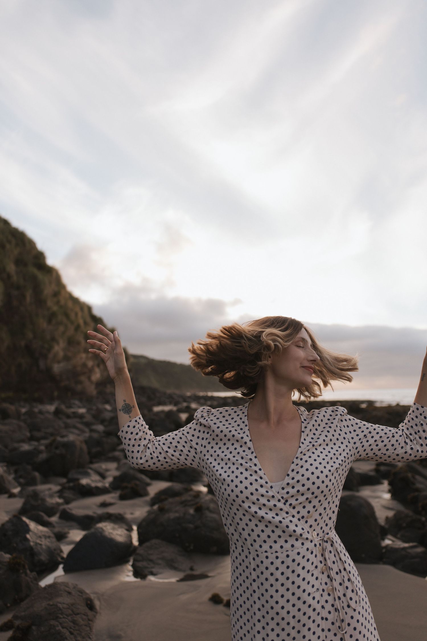 Woman in polka dot dress with arms outstretched on rocky beach, hair blowing in wind at sunset in New Zealand.