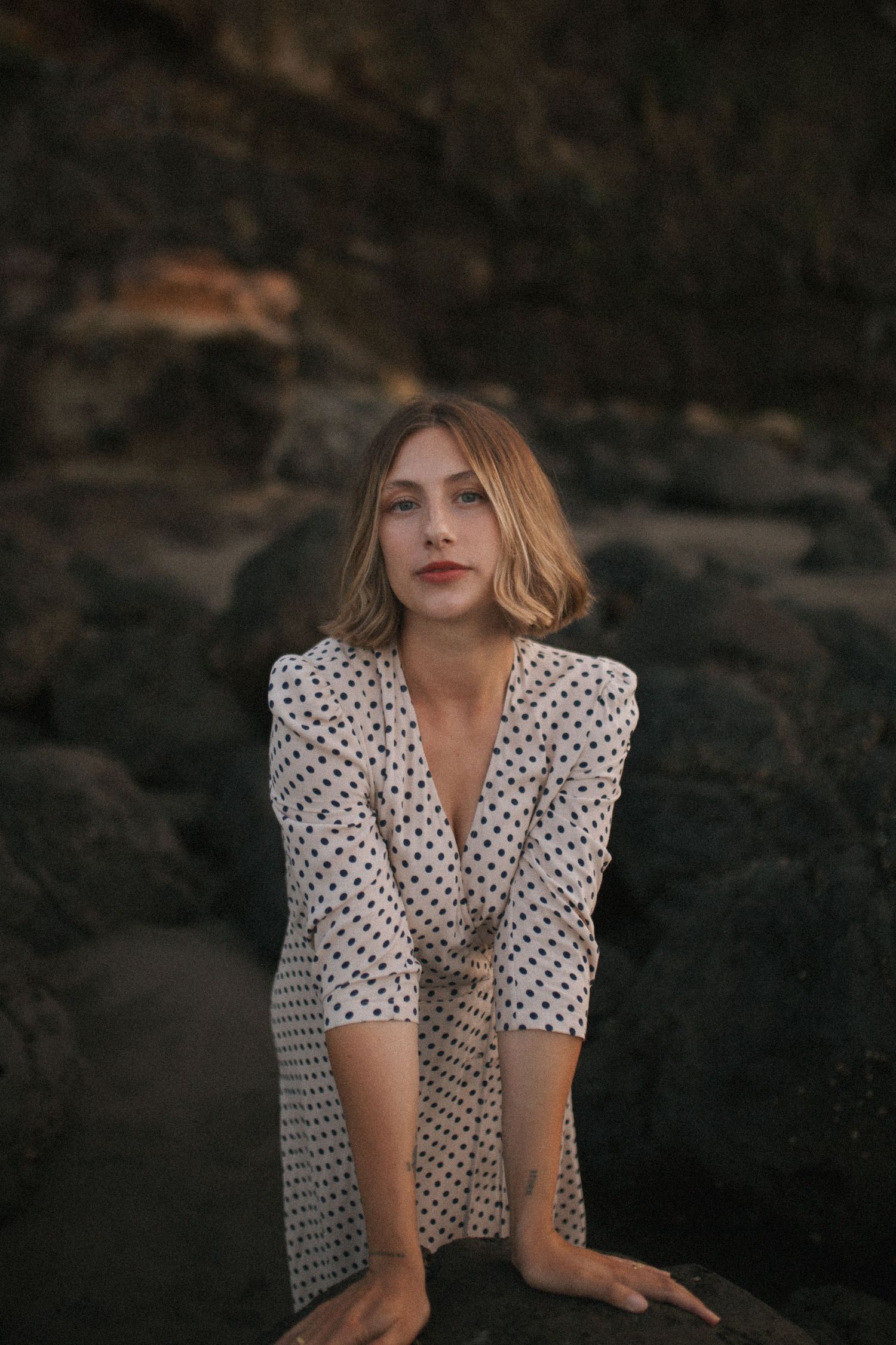 Young woman in white polka dot wrap dress posing against dark rocky coastal background in New Zealand.