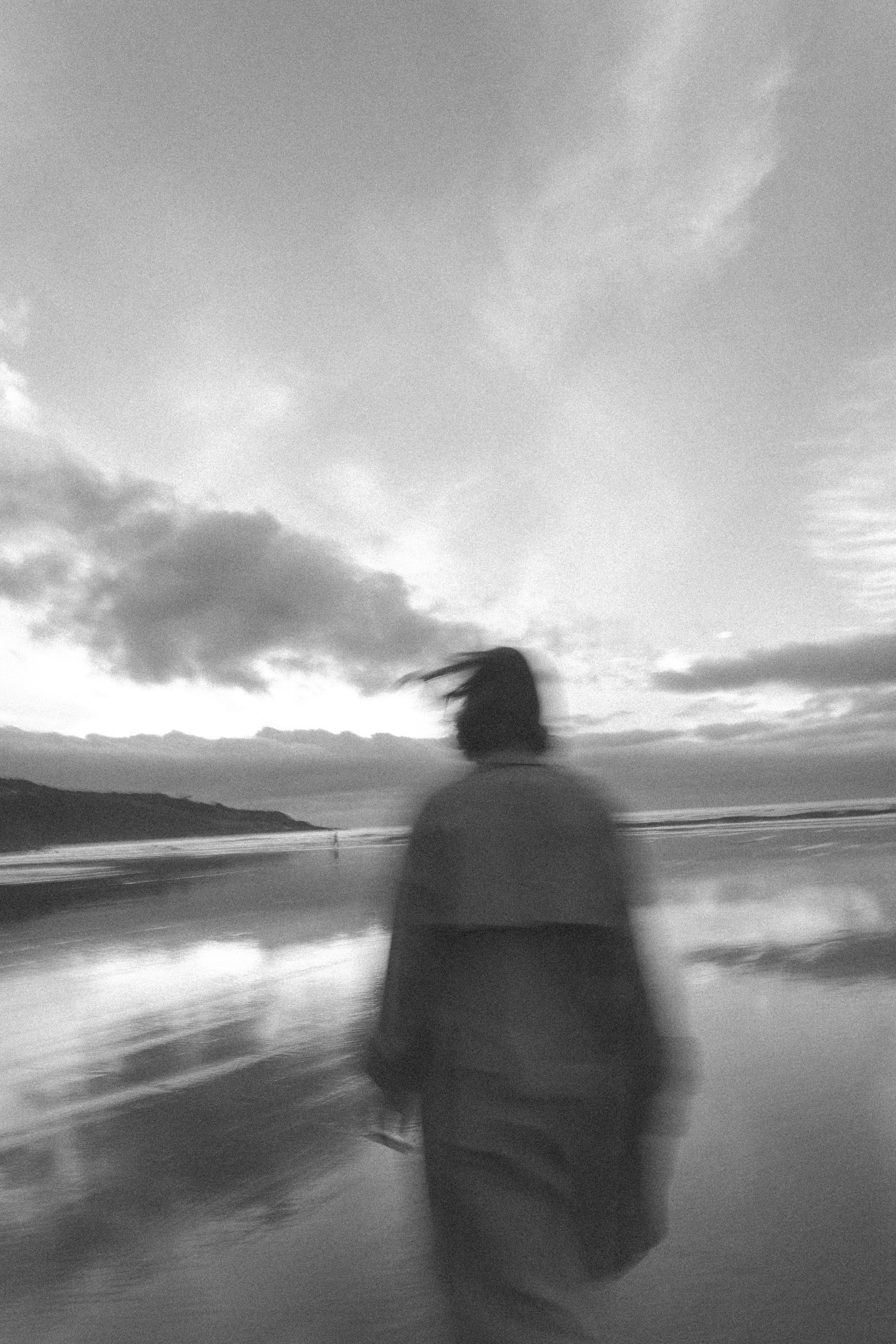 Blurred black and white silhouette of woman walking on reflective wet beach at sunset in New Zealand.