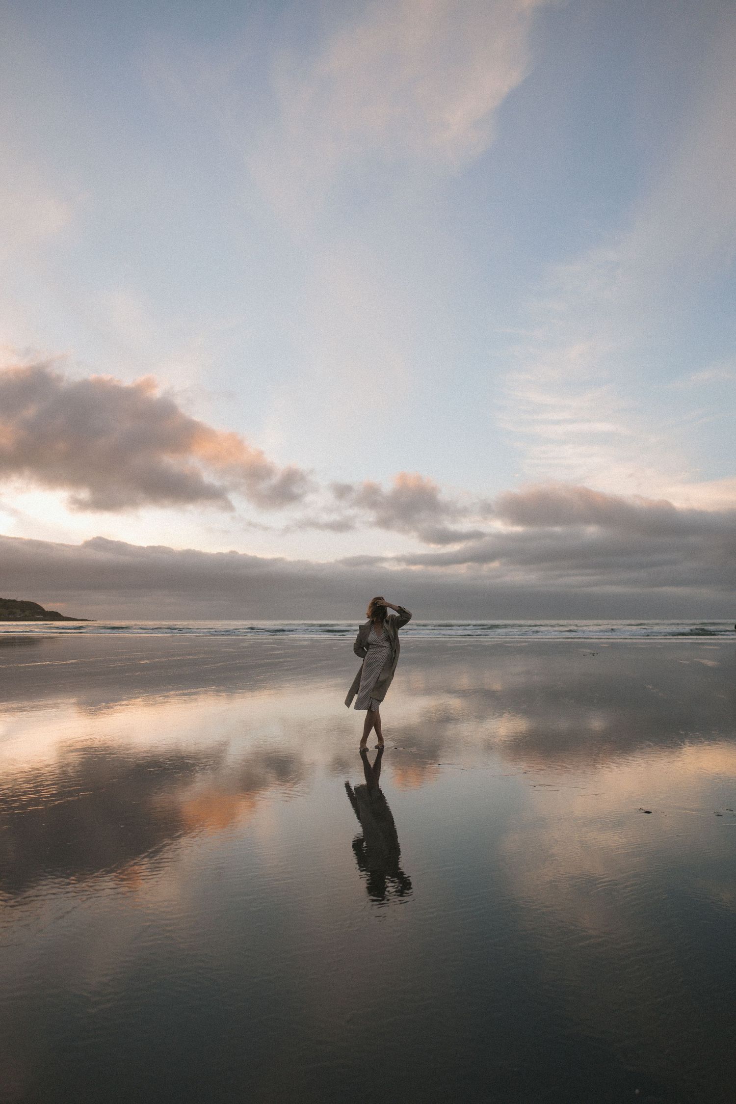 Woman in dress standing on reflective wet beach at sunset with dramatic cloud reflections in New Zealand.