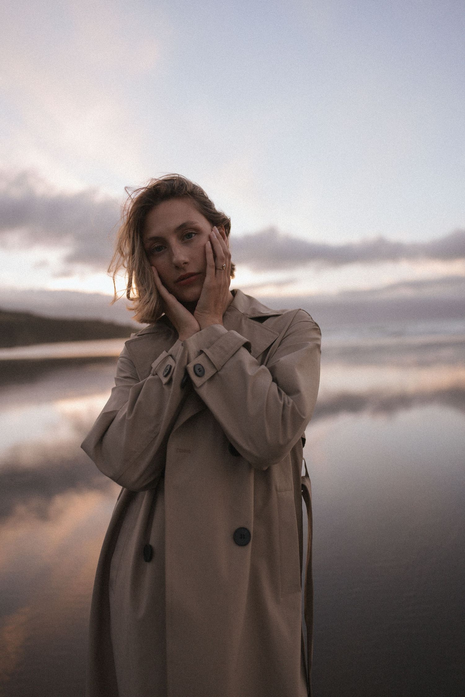 Young woman in beige trench coat holding face with both hands on reflective sunset beach in New Zealand.