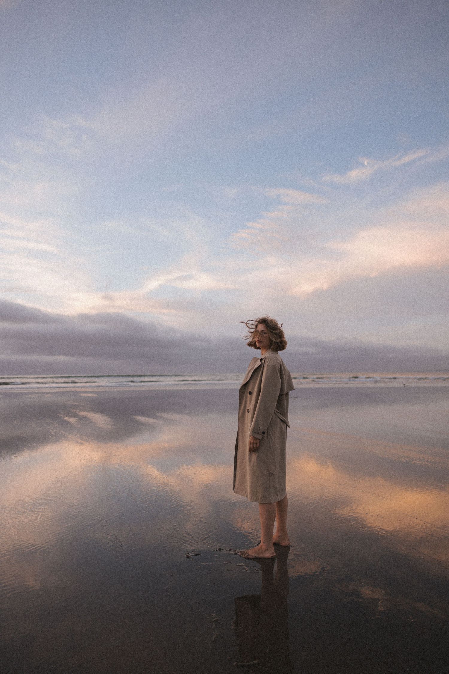 Woman in beige trench coat standing barefoot on reflective wet beach at pastel sunset in New Zealand.