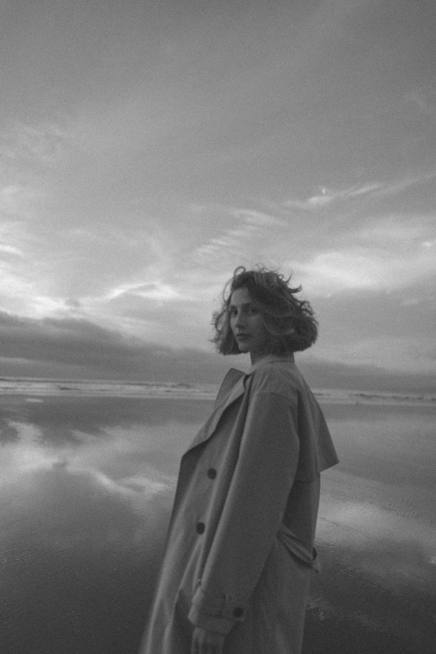 Black and white portrait of a woman in a trench coat standing on a reflective beach under dramatic cloudy skies in New Zealand.