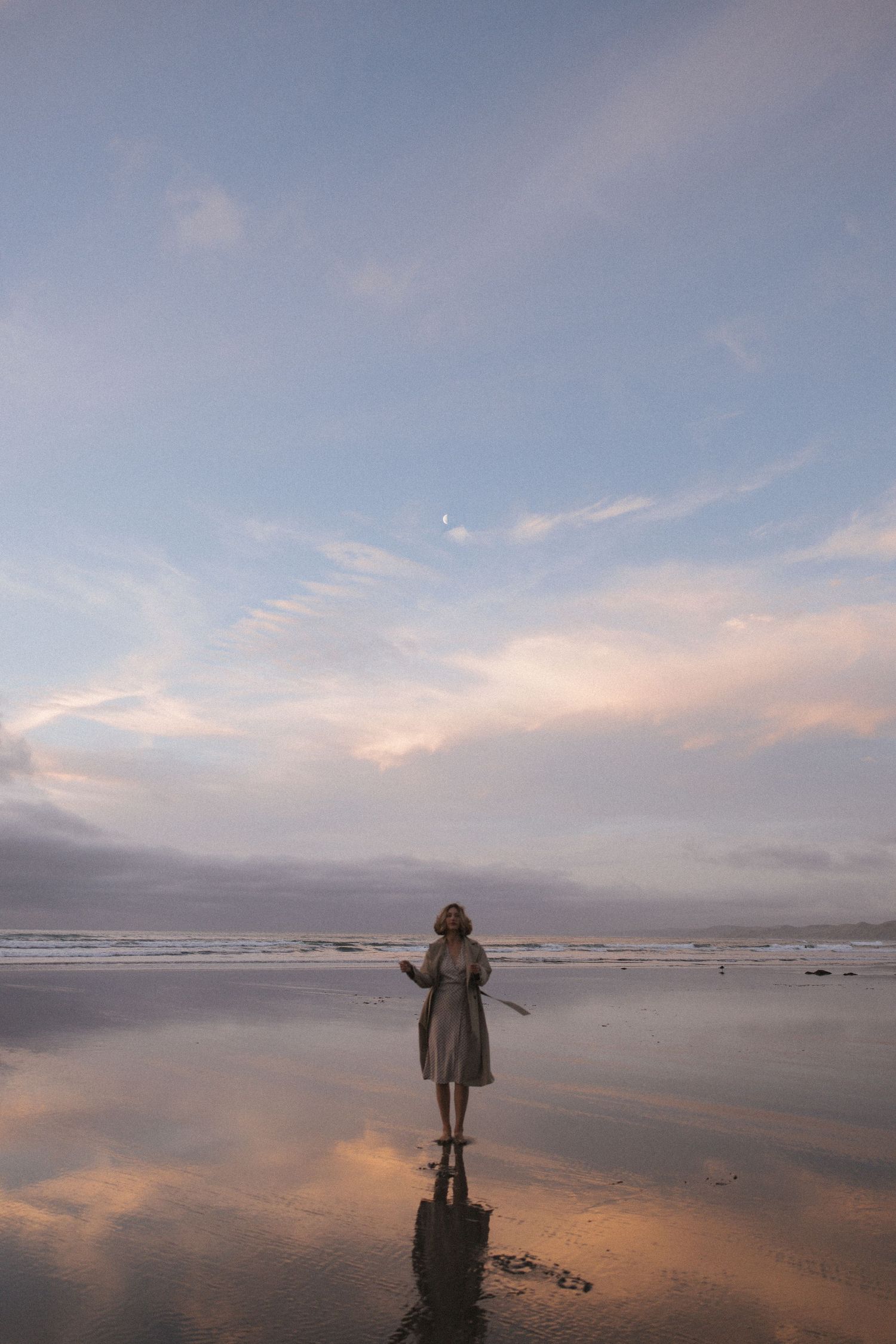 Woman in a coat standing alone on a wet reflective beach at sunset with a crescent moon visible in New Zealand.