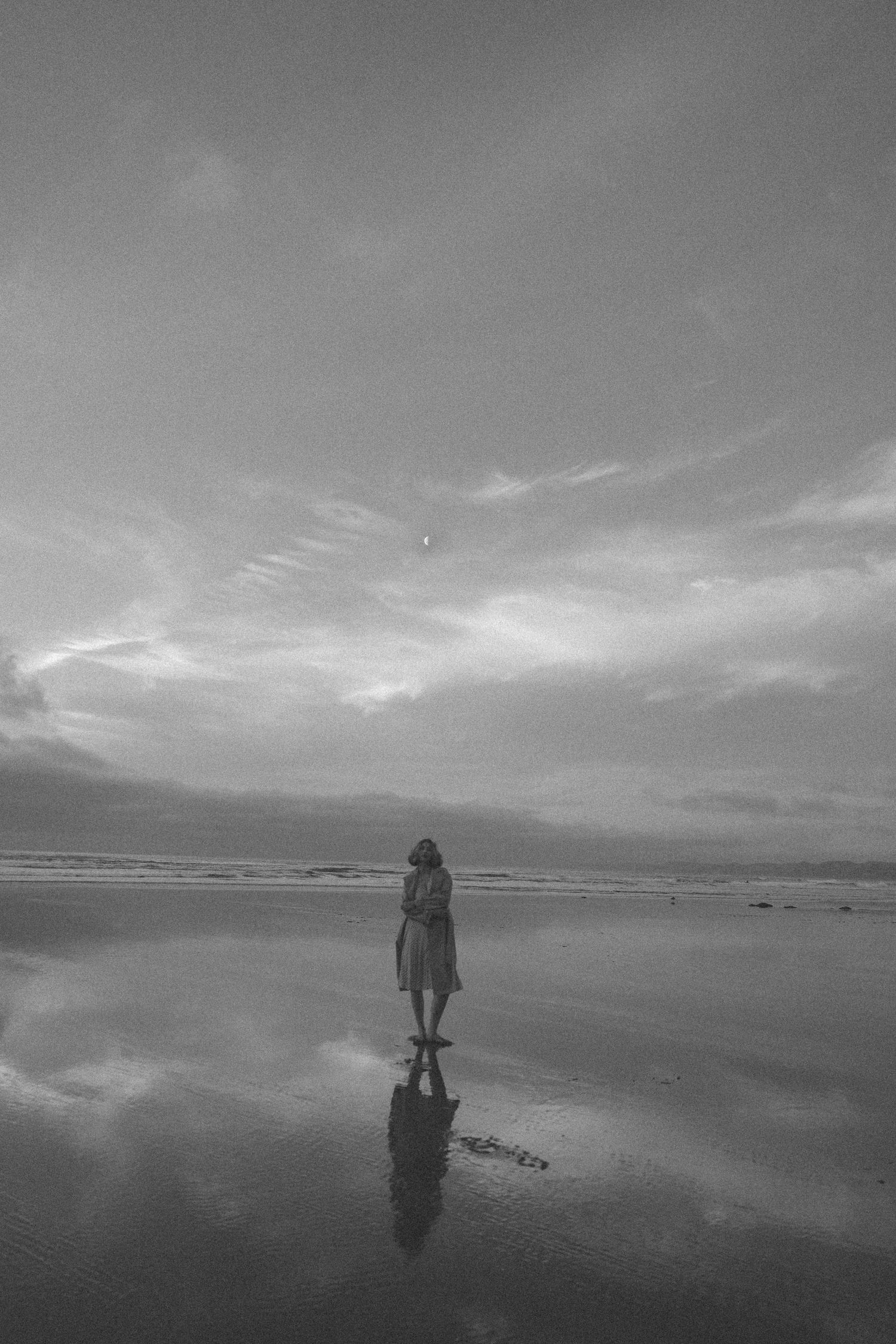 Black and white silhouette of lone woman standing on reflective wet beach under dramatic sky in New Zealand.