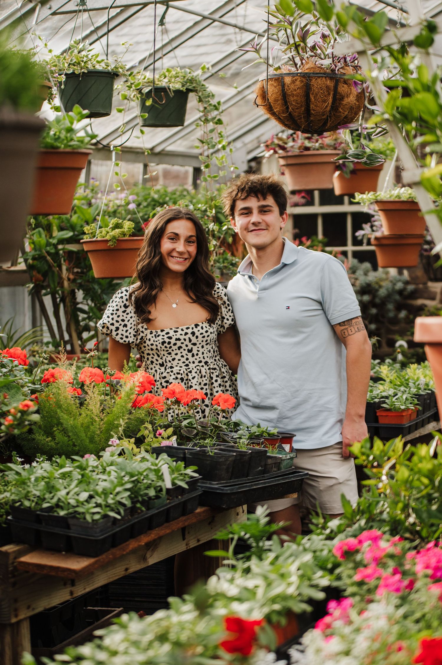 Two people stand together in a greenhouse filled with colorful flowers and hanging plants on display.