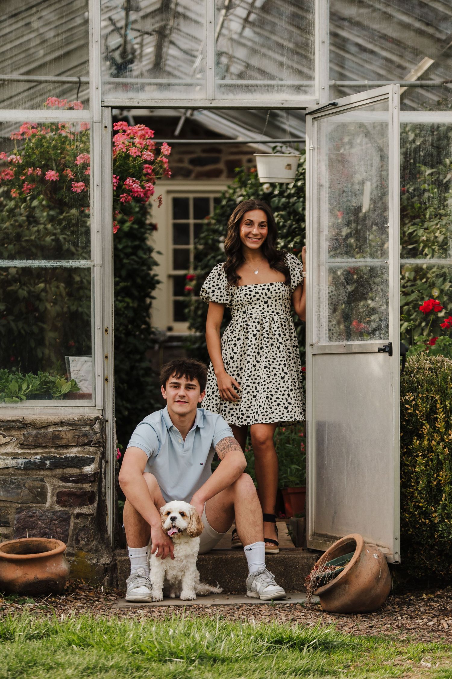 A couple sits with their small white dog in front of a greenhouse surrounded by blooming roses and stone walls.