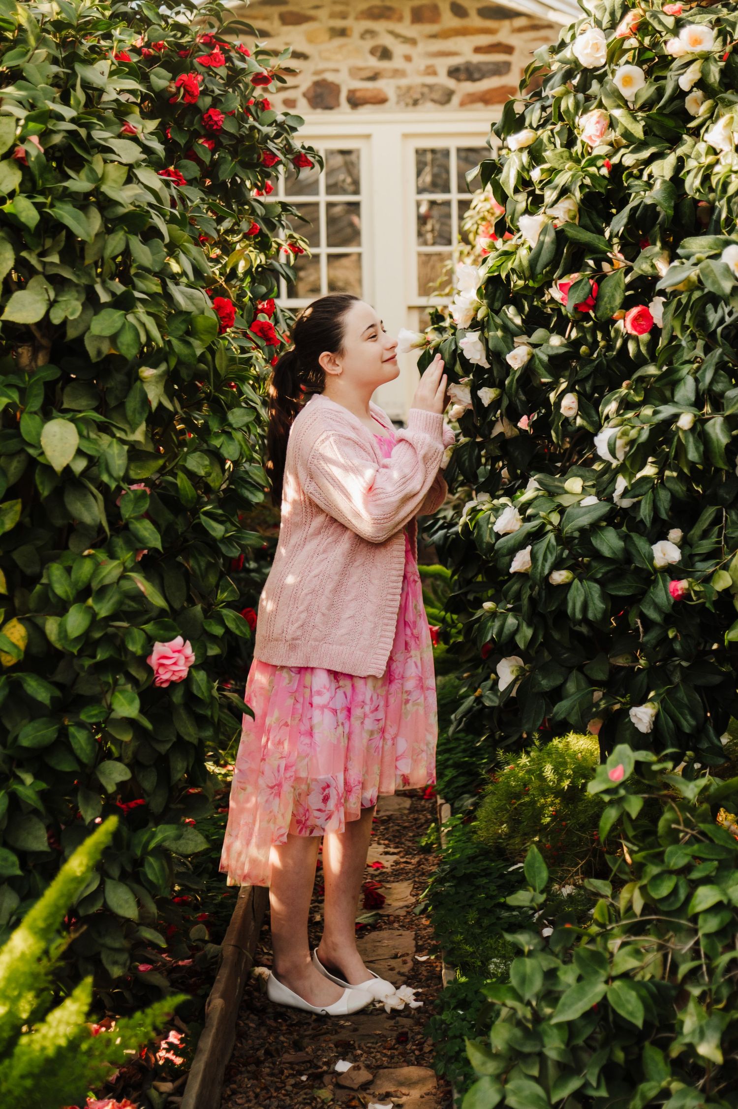 A pink dress against a stone wall backdrop with lush green foliage and red flowers in a garden setting.