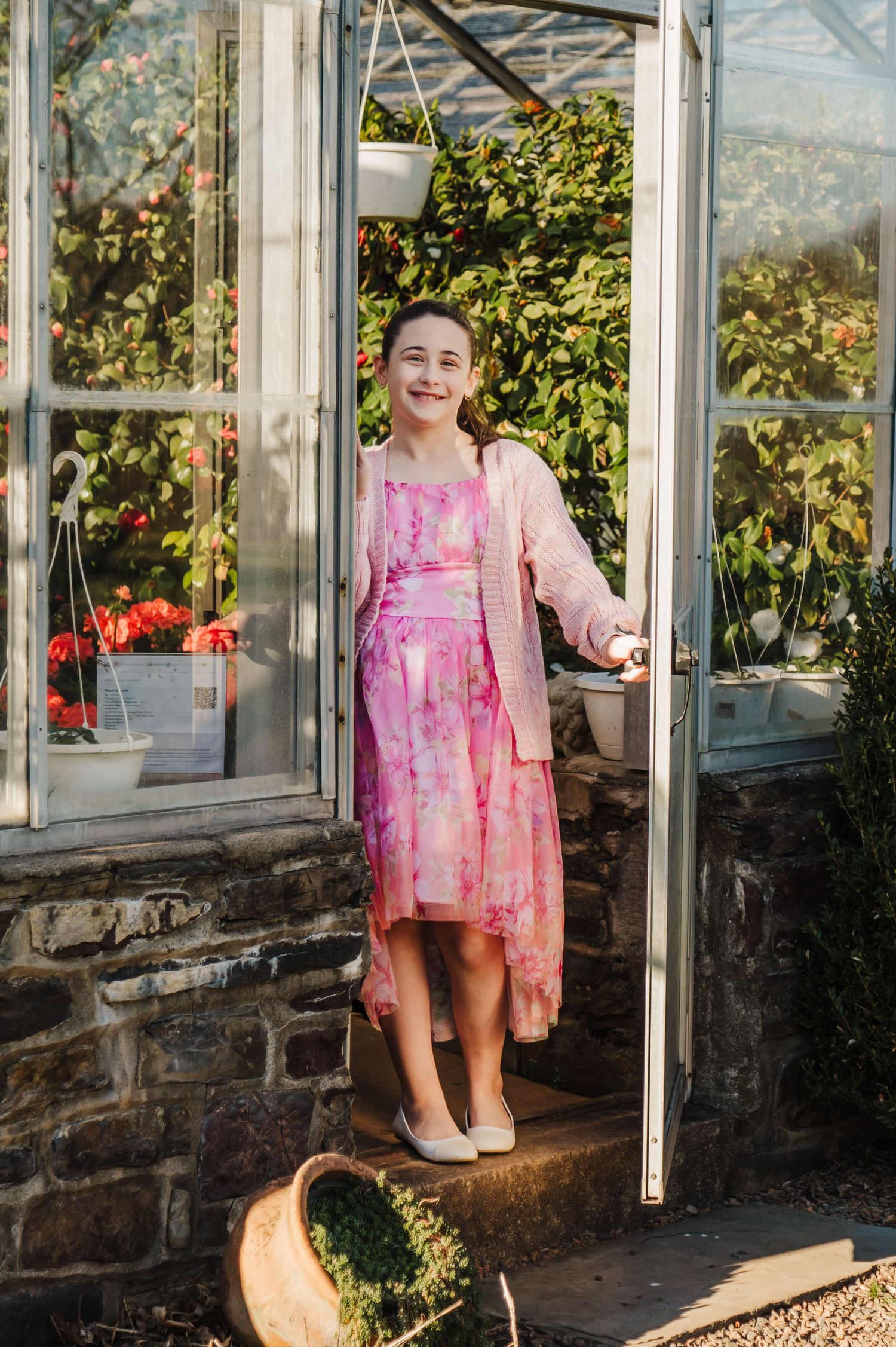 Person in pink floral dress stands outside a greenhouse with glass panels and stone walls in warm sunlight.