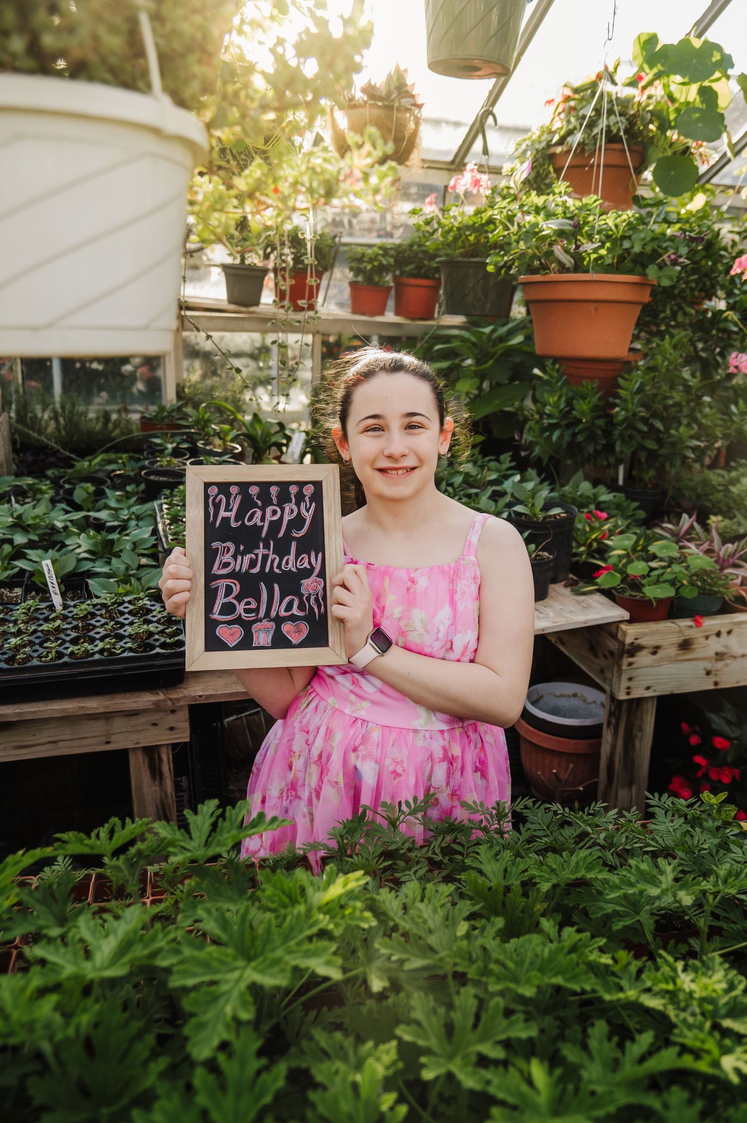 A pink dress-clad figure sits in a lush greenhouse garden setting holding a chalkboard sign.