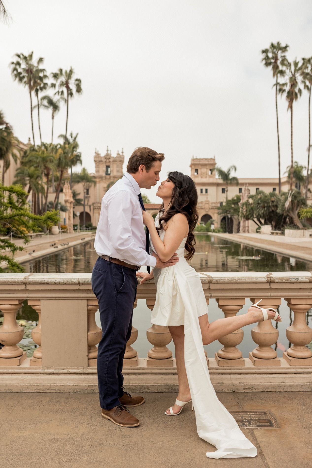 A romantic couple shares a kiss while striking playful poses on a balcony with palm trees and Spanish architecture in the background.