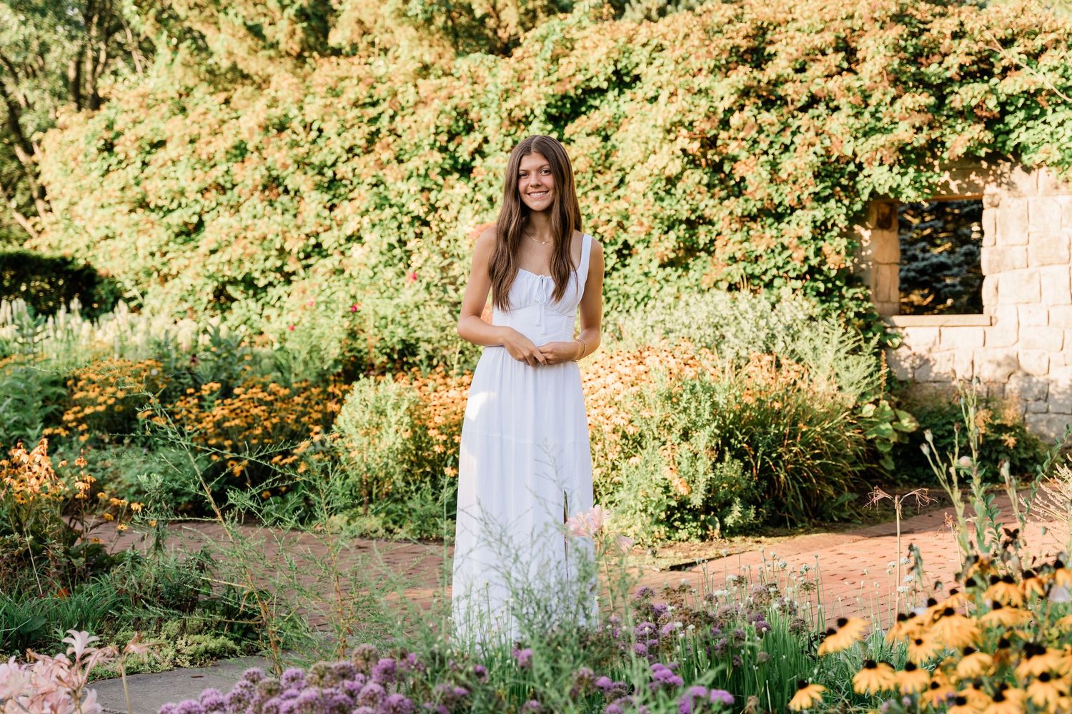 A person in a white lace dress poses in a lush garden with blooming orange and purple flowers.