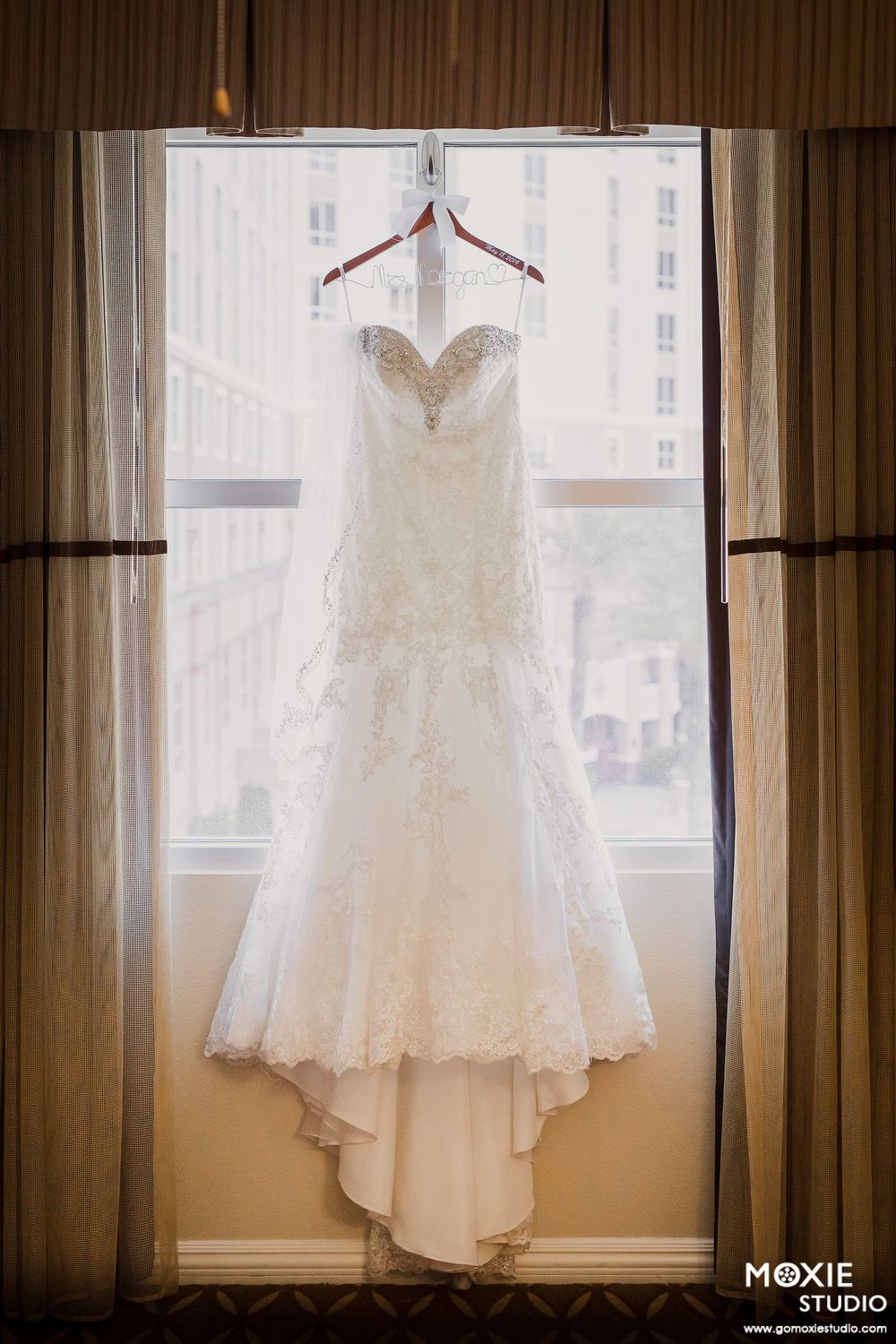 A beautiful white lace wedding dress hangs by a window in natural light, showcasing its delicate details and flowing train.