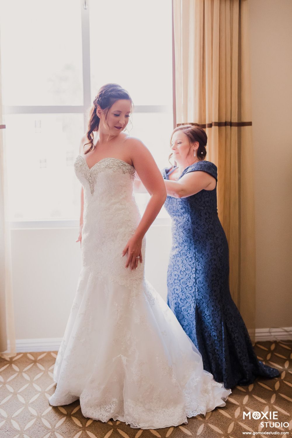 A strapless lace wedding gown is being fitted while someone in a navy blue dress assists with adjustments.