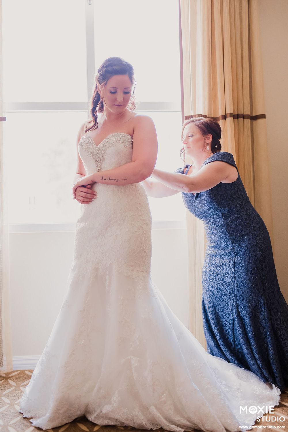 A woman in a white lace wedding gown gets help with final adjustments from someone wearing a blue dress.