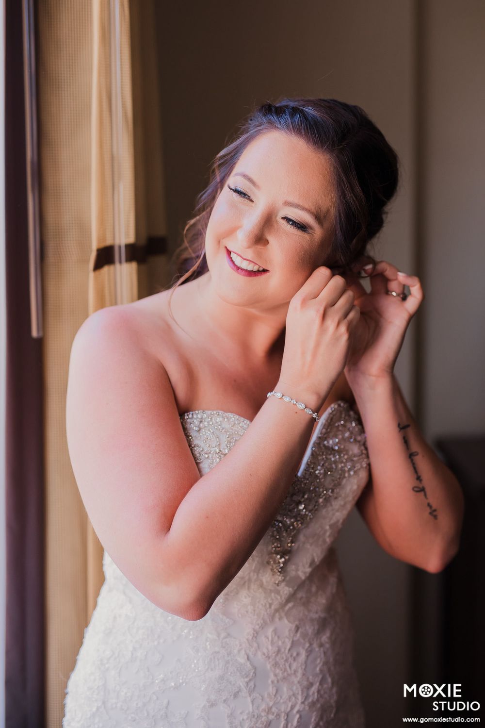 Close up of bride wearing a sparkling strapless wedding dress while putting on earrings near a window.