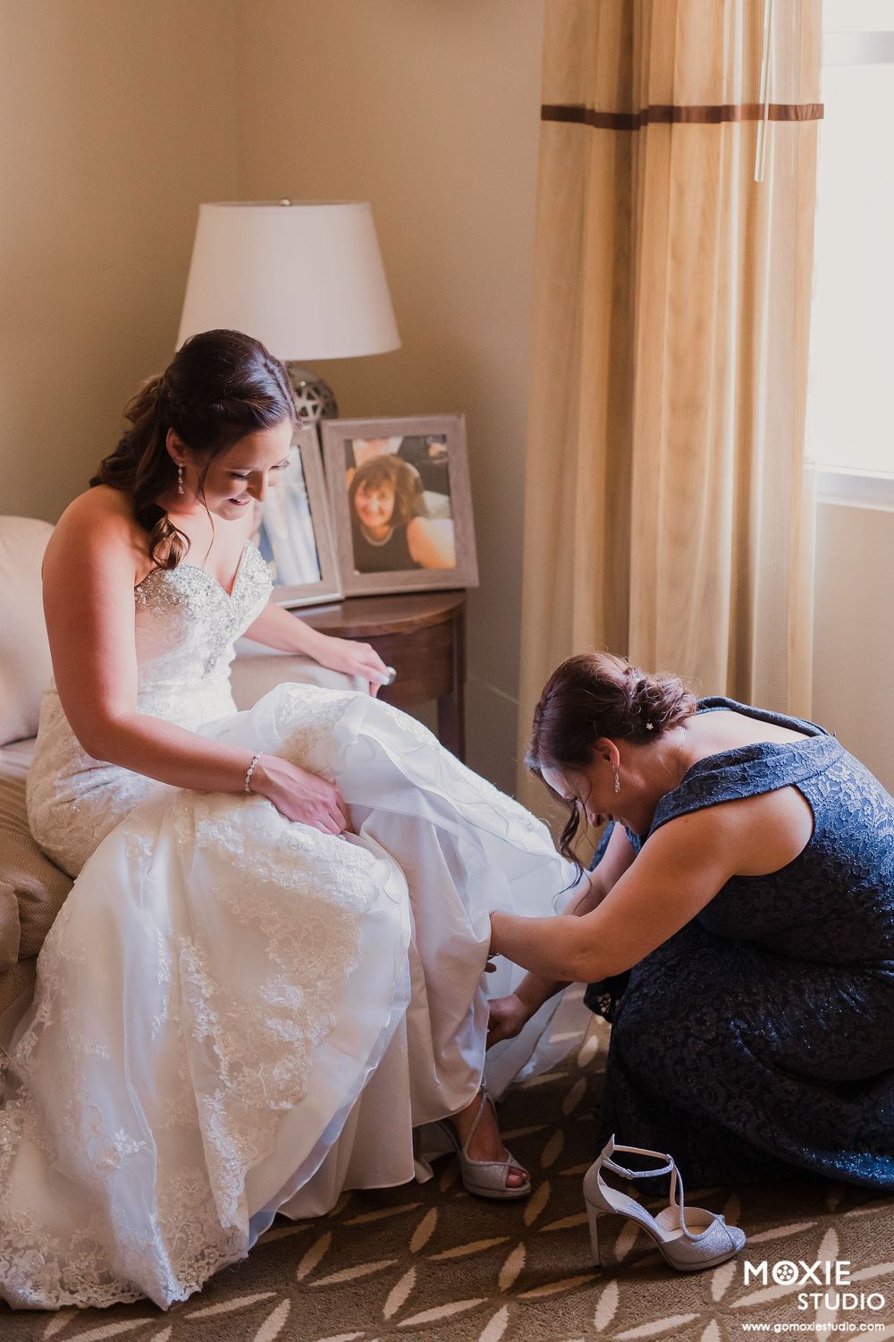 A bride in a white lace wedding dress gets help putting on her shoes during wedding preparations.