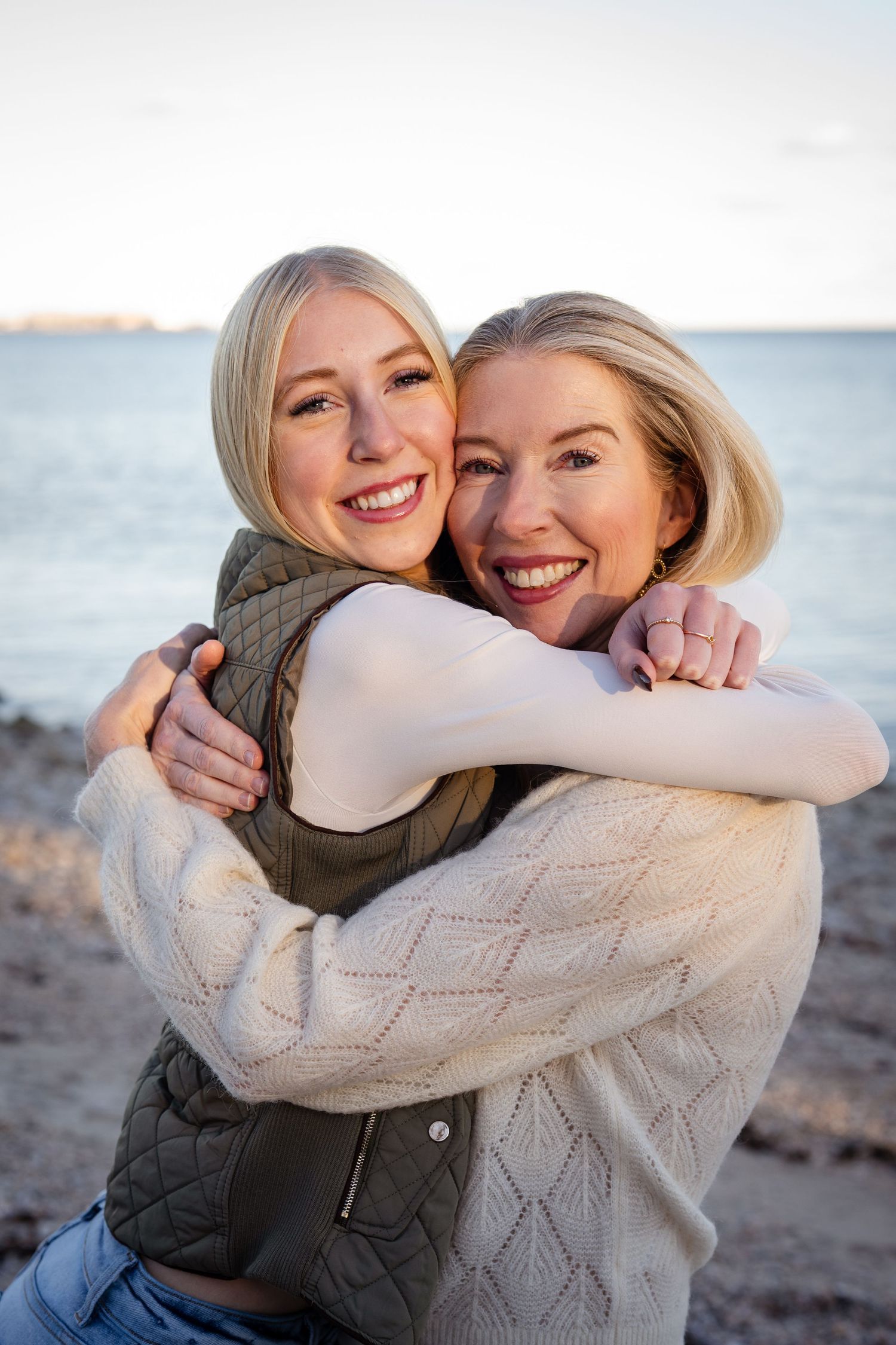 A mother and her teen daughter embracing and laughing together on a beach during sunset.