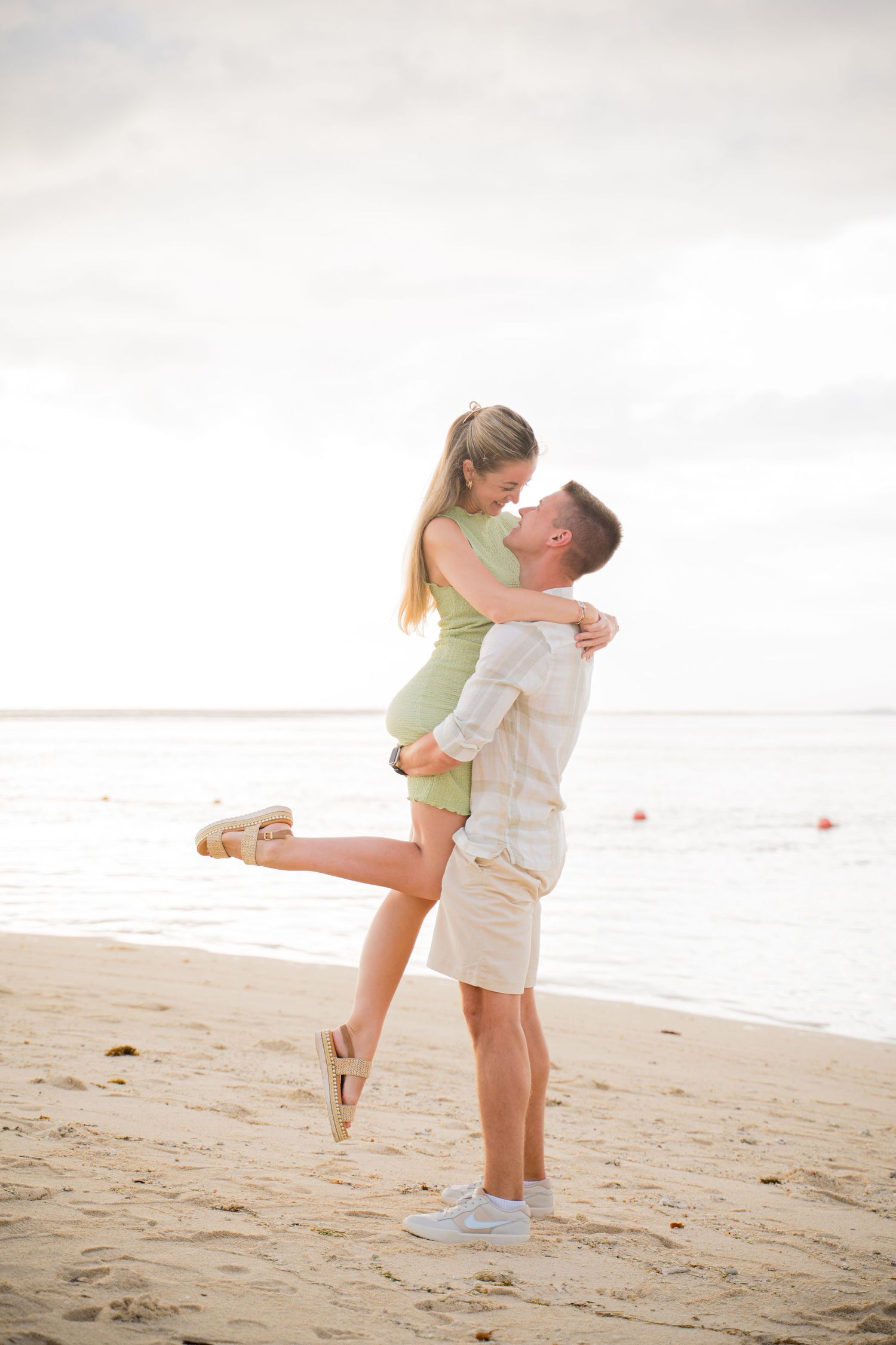 A romantic couple embraces on a sunny beach, with one person lifted in the air against a bright cloudy sky.