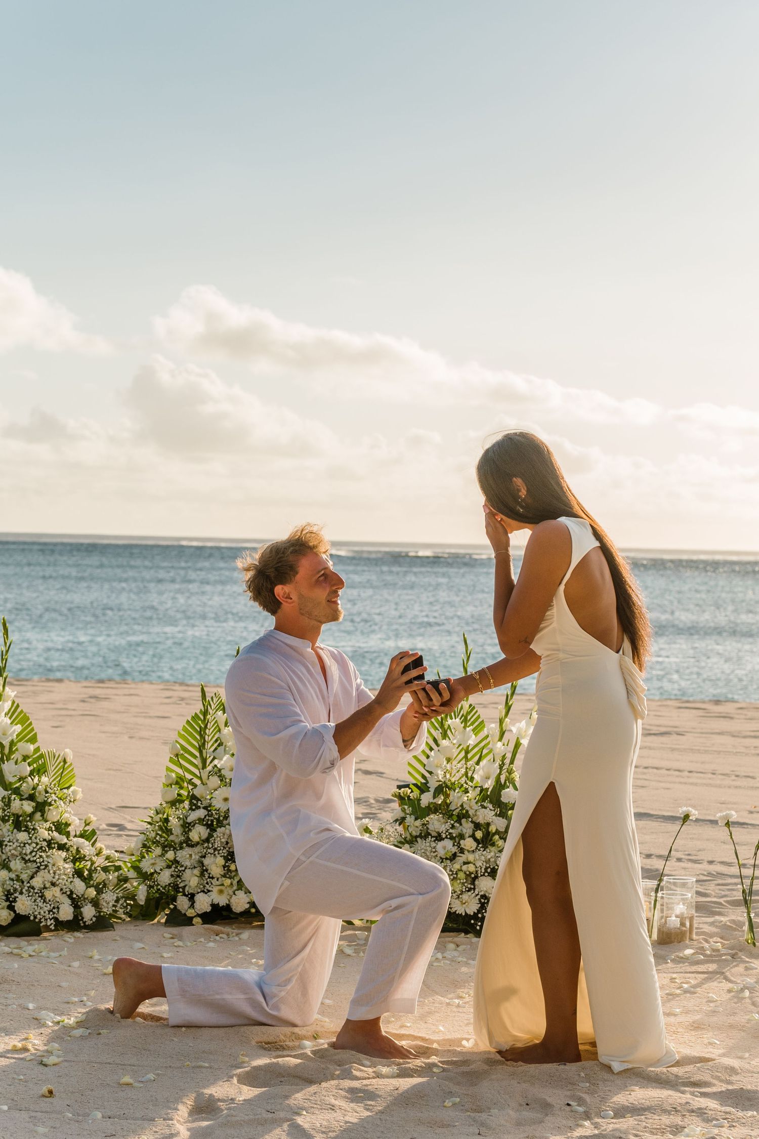 A romantic couple embraces on a sunny beach, with one person lifted in the air against a bright cloudy sky.