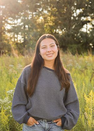 A person in a striped sweater smiles while standing in a sunlit meadow with tall grass.