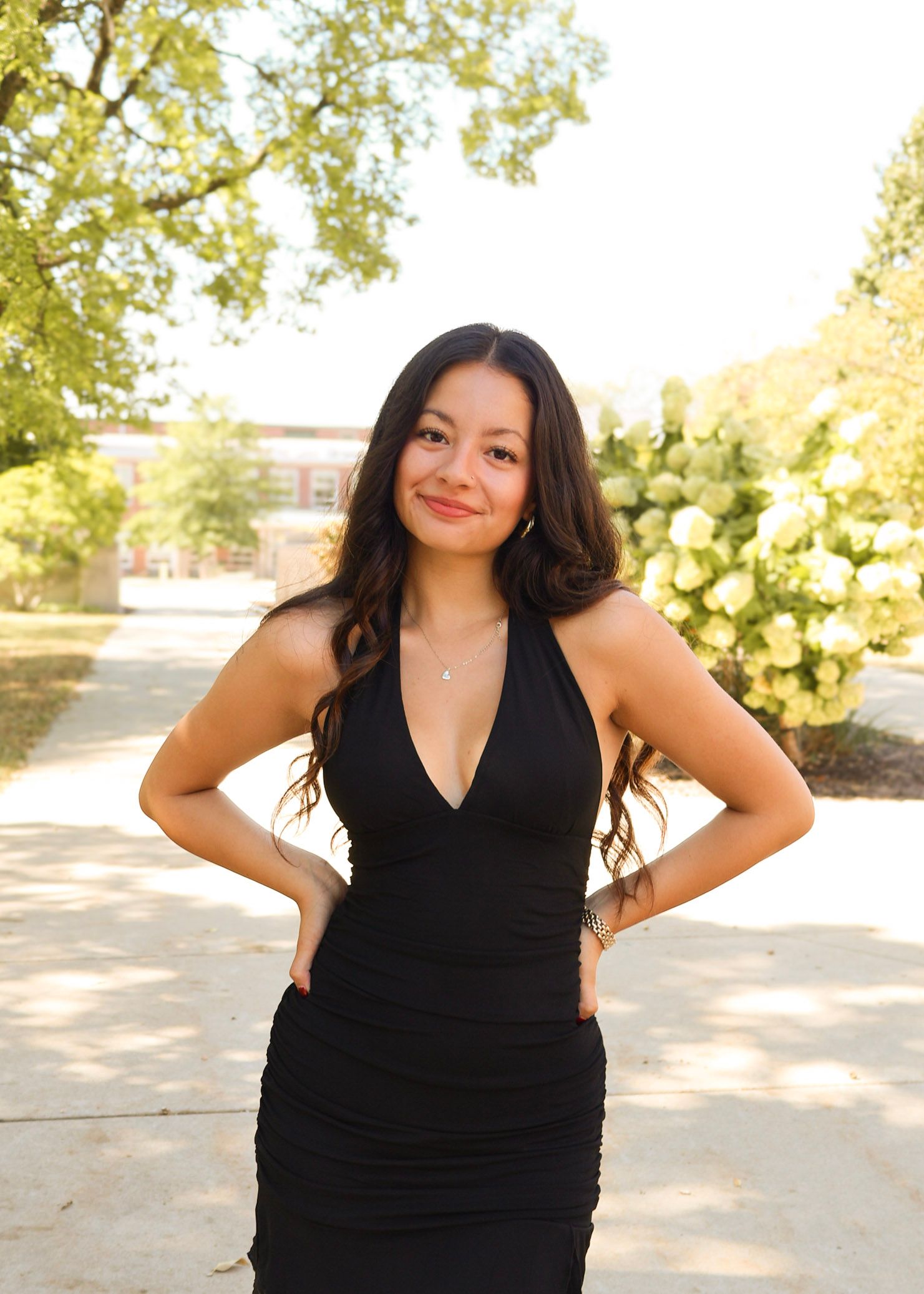 Elegant poses in a black v-neck dress against a sunlit outdoor setting.