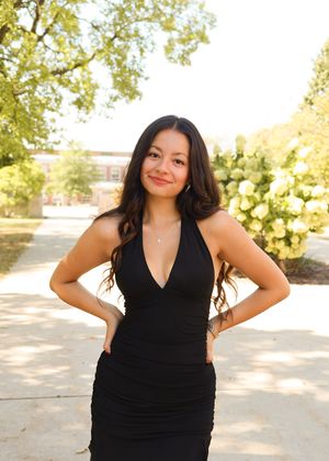 Elegant poses in a black v-neck dress against a sunlit outdoor setting.