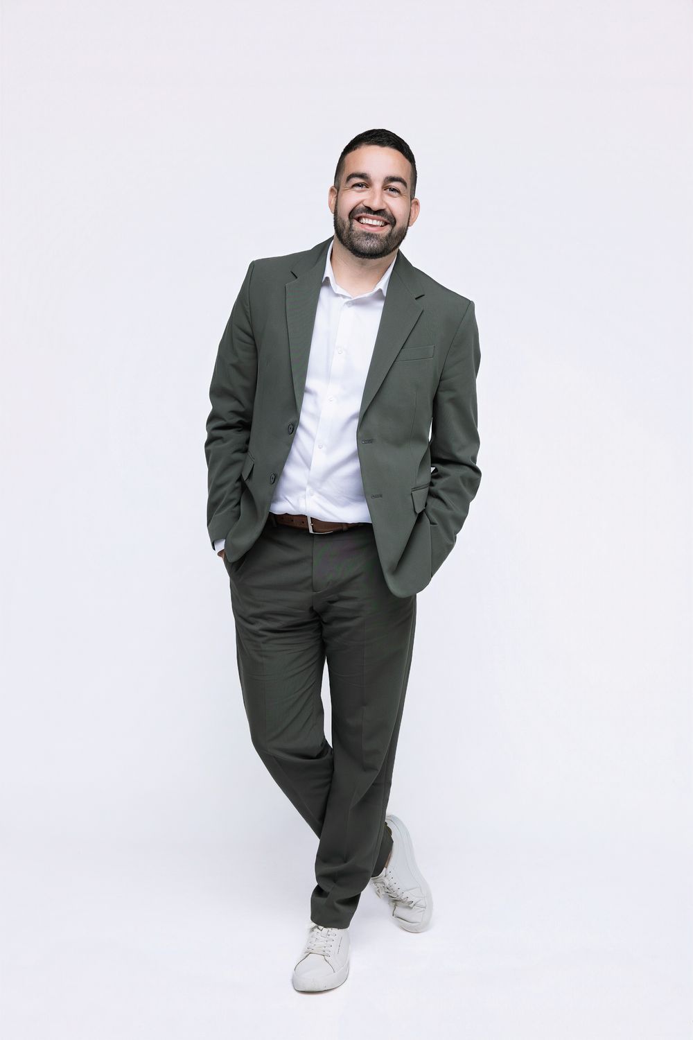 A person wearing a dark green suit and white sneakers poses confidently against a white background.