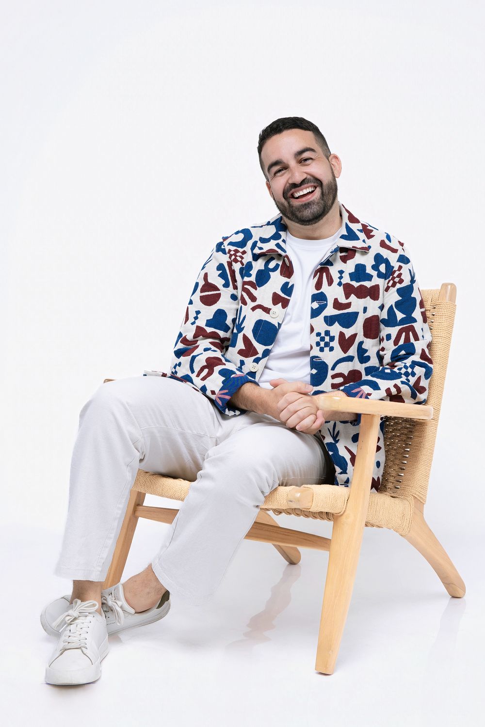 Person in white pants and patterned shirt relaxes on wooden lounge chair against white background.