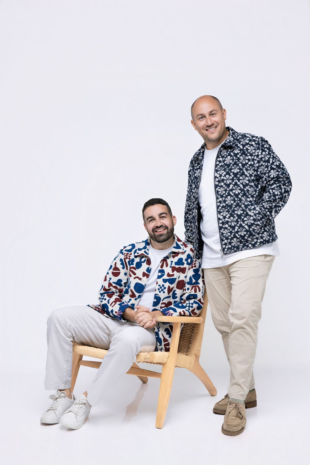 Two people in coordinating patterned sweaters sit and pose together on a wooden chair against a white backdrop.