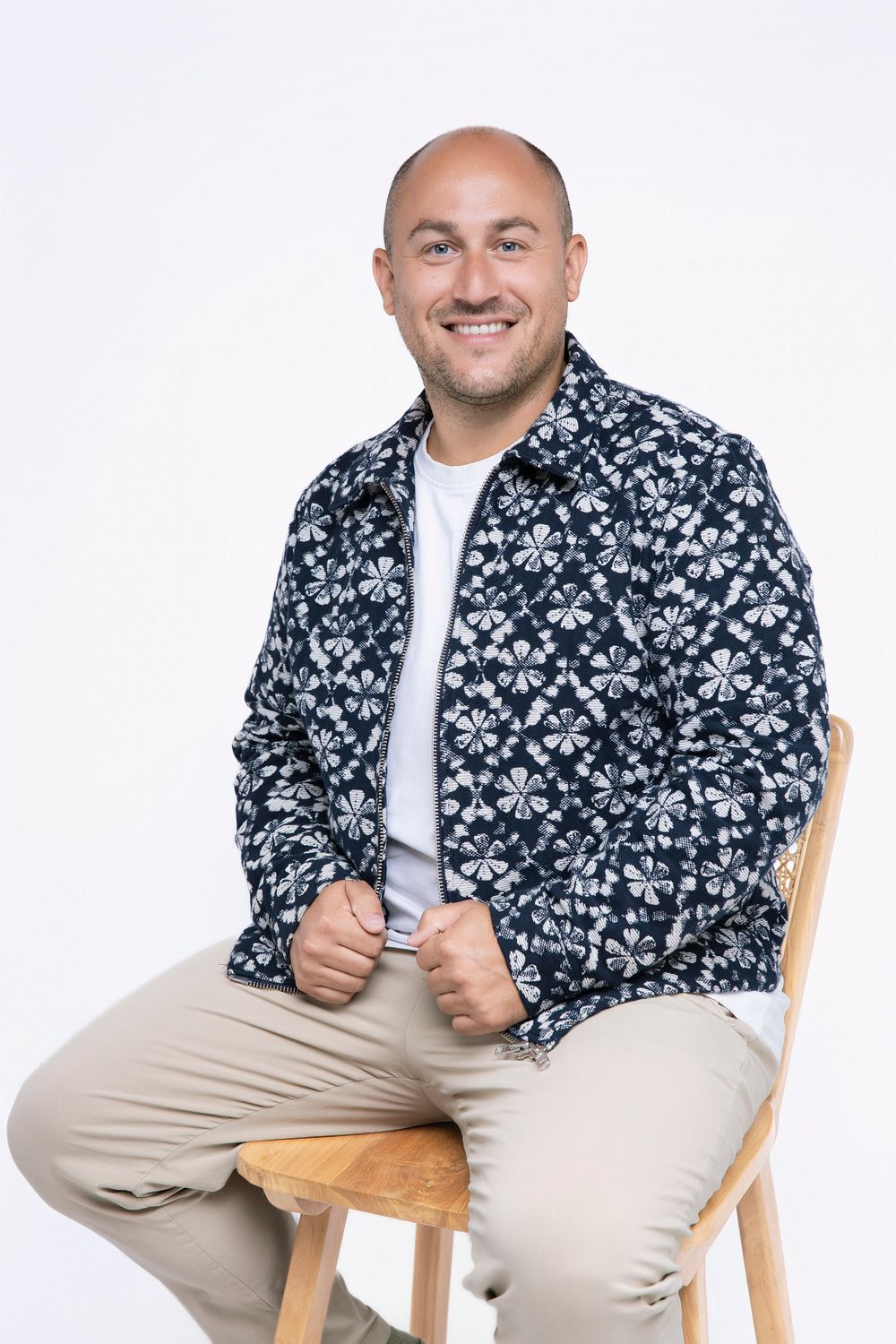 Person wearing a patterned black and white jacket sits smiling on a wooden chair against white backdrop.