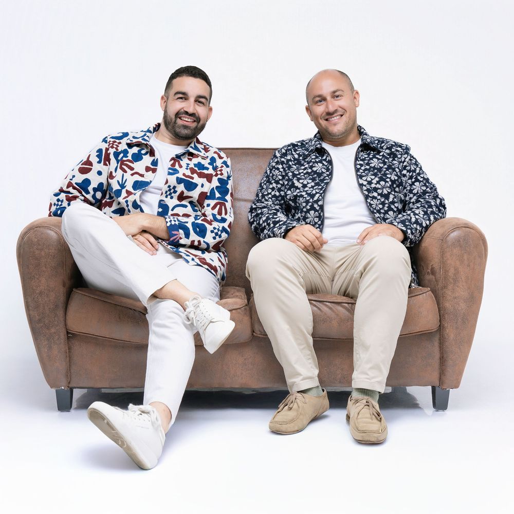 Two people wearing matching patterned shirts pose casually on a brown leather couch in a studio setting.
