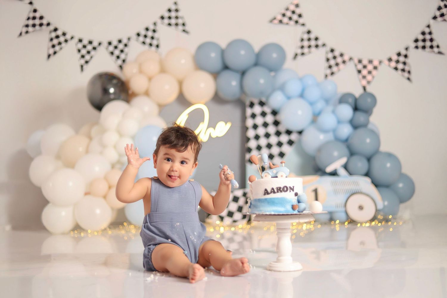 A baby's first birthday cake smash photo session with blue and white balloon decorations and checkered racing flags.