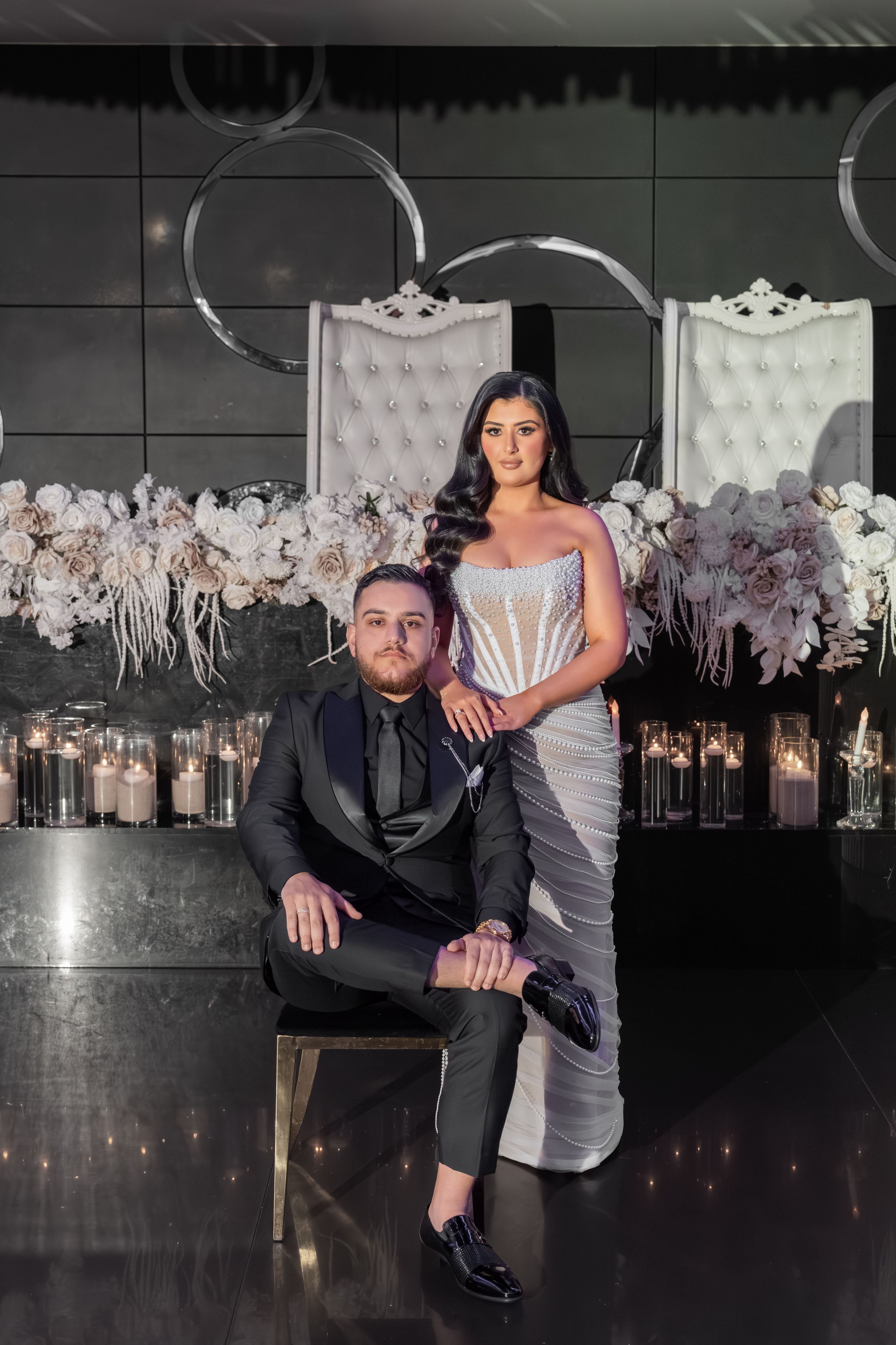 Elegant couple poses in formal attire against a backdrop of white floral arrangements and ornate chairs in a dark venue.