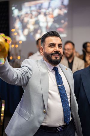 A person in a light gray suit and blue tie smiles and gestures during what appears to be a celebratory event.