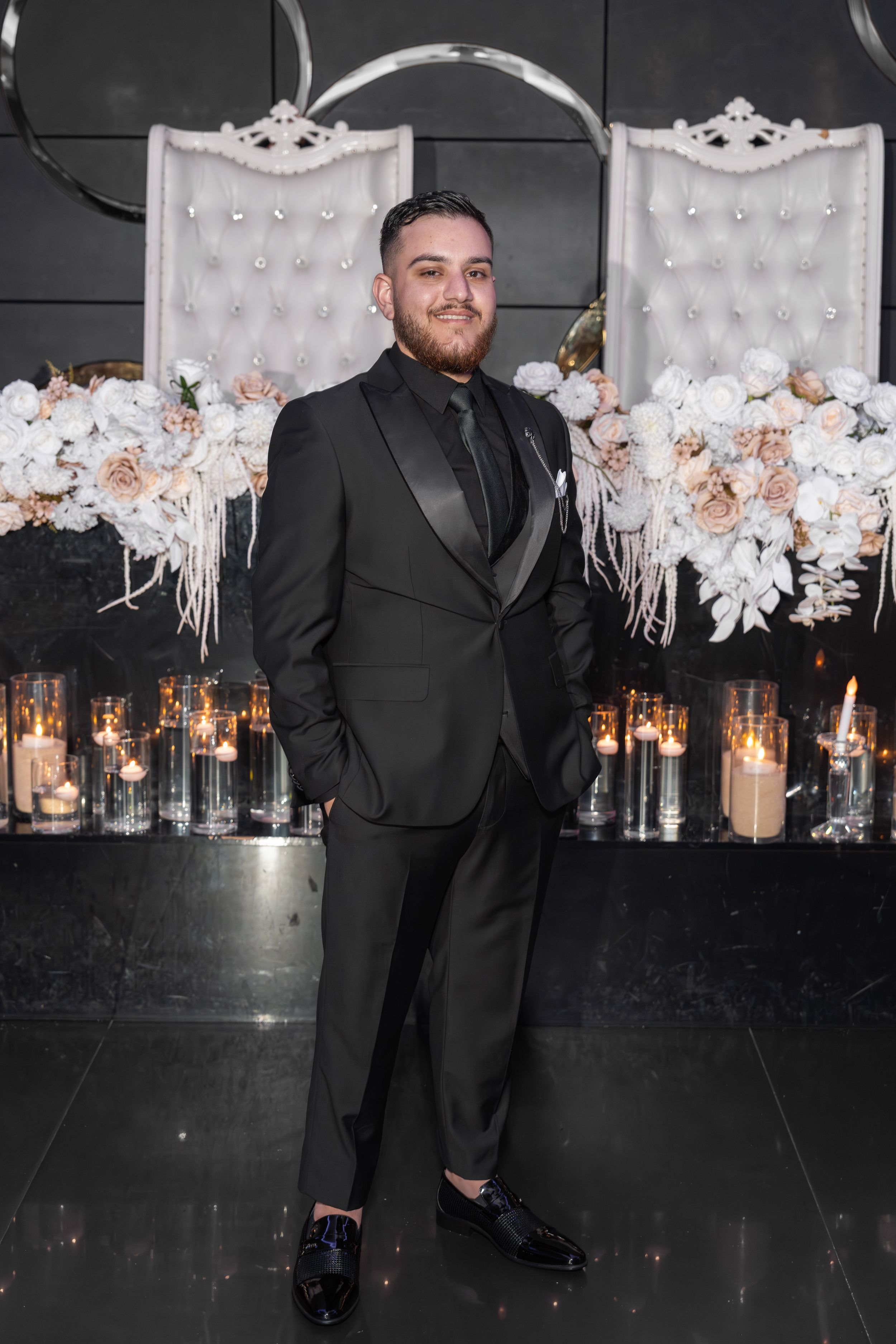 A person in a black suit poses at a formal event with elegant white floral decorations and candlelit decor in the background.