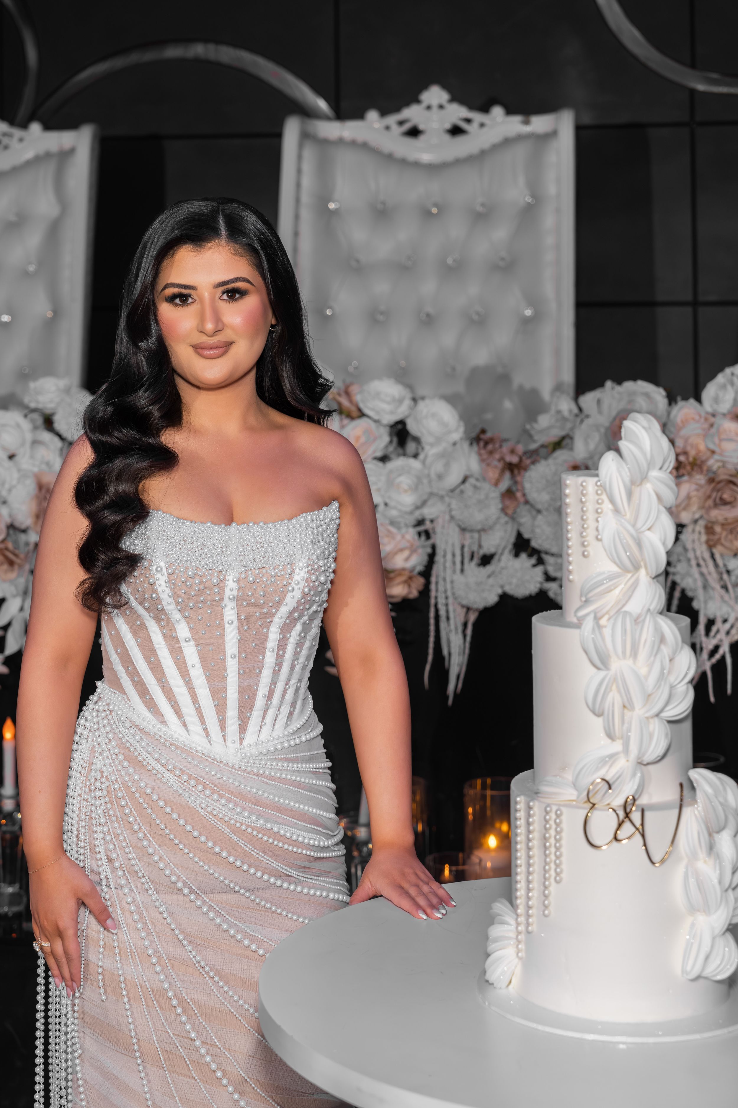 A person in a white beaded gown poses next to an elegant white tiered cake at an event.