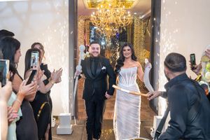 A group of elegantly dressed guests enjoy a formal event in a luxurious ballroom with a crystal chandelier.