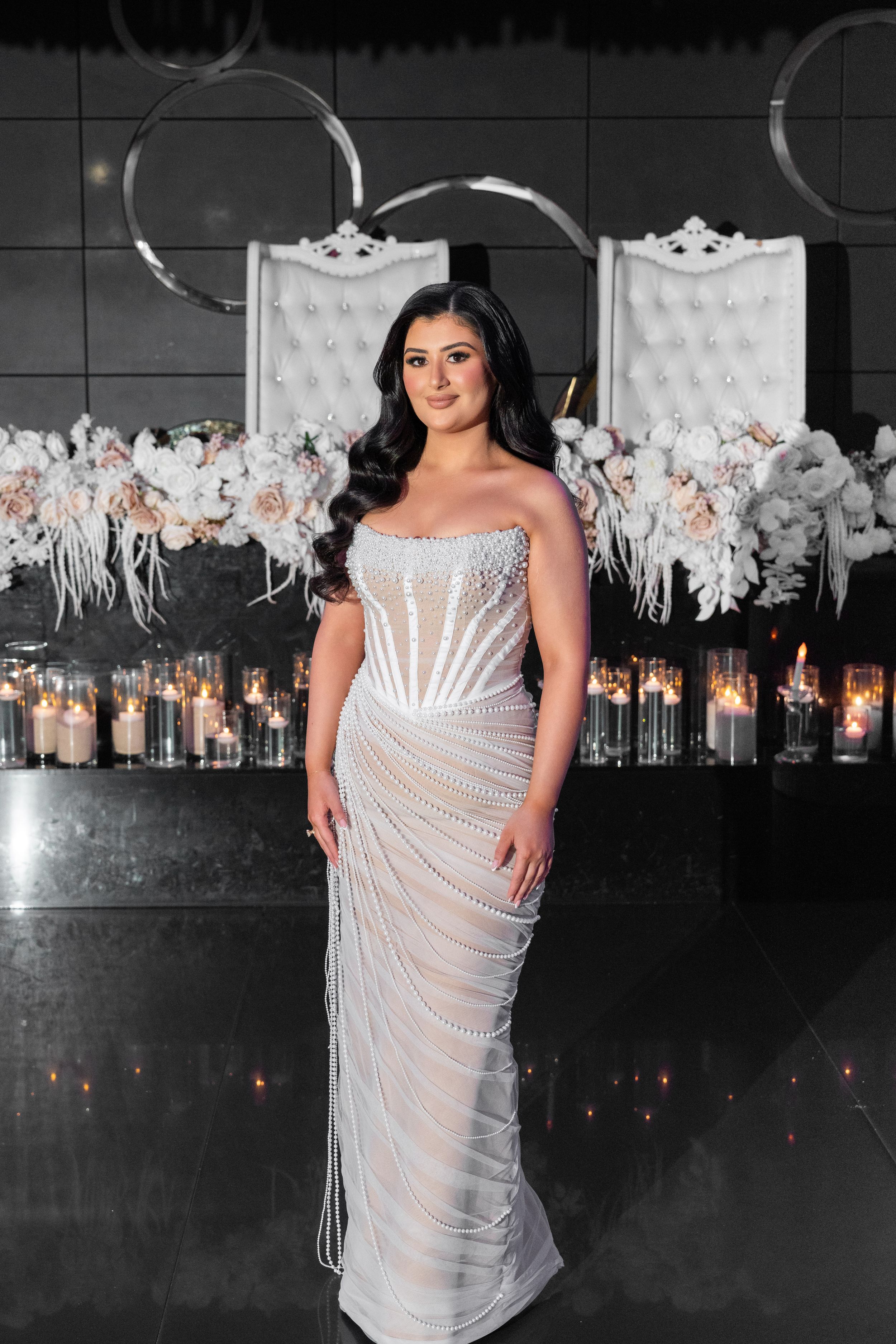 A plus-size model poses in a glamorous white beaded gown against a dark background with hanging floral decorations.