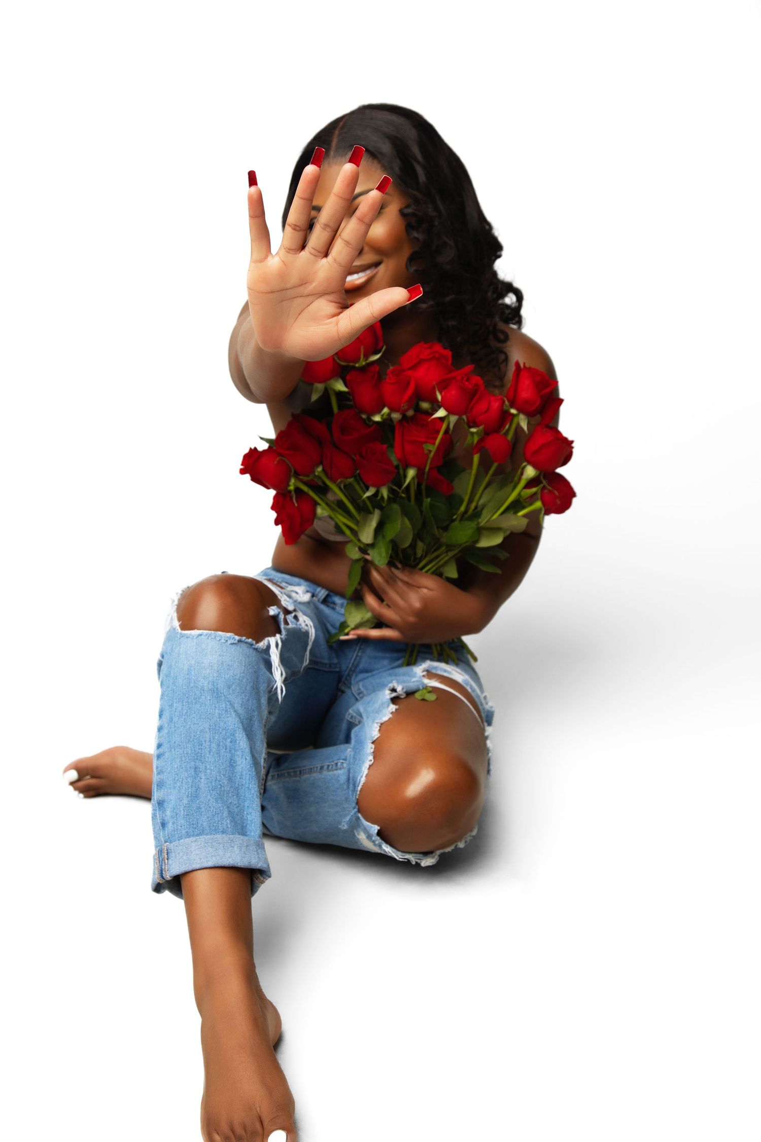 Woman sitting on floor holding red roses while blocking her face with her hand, wearing ripped jeans.