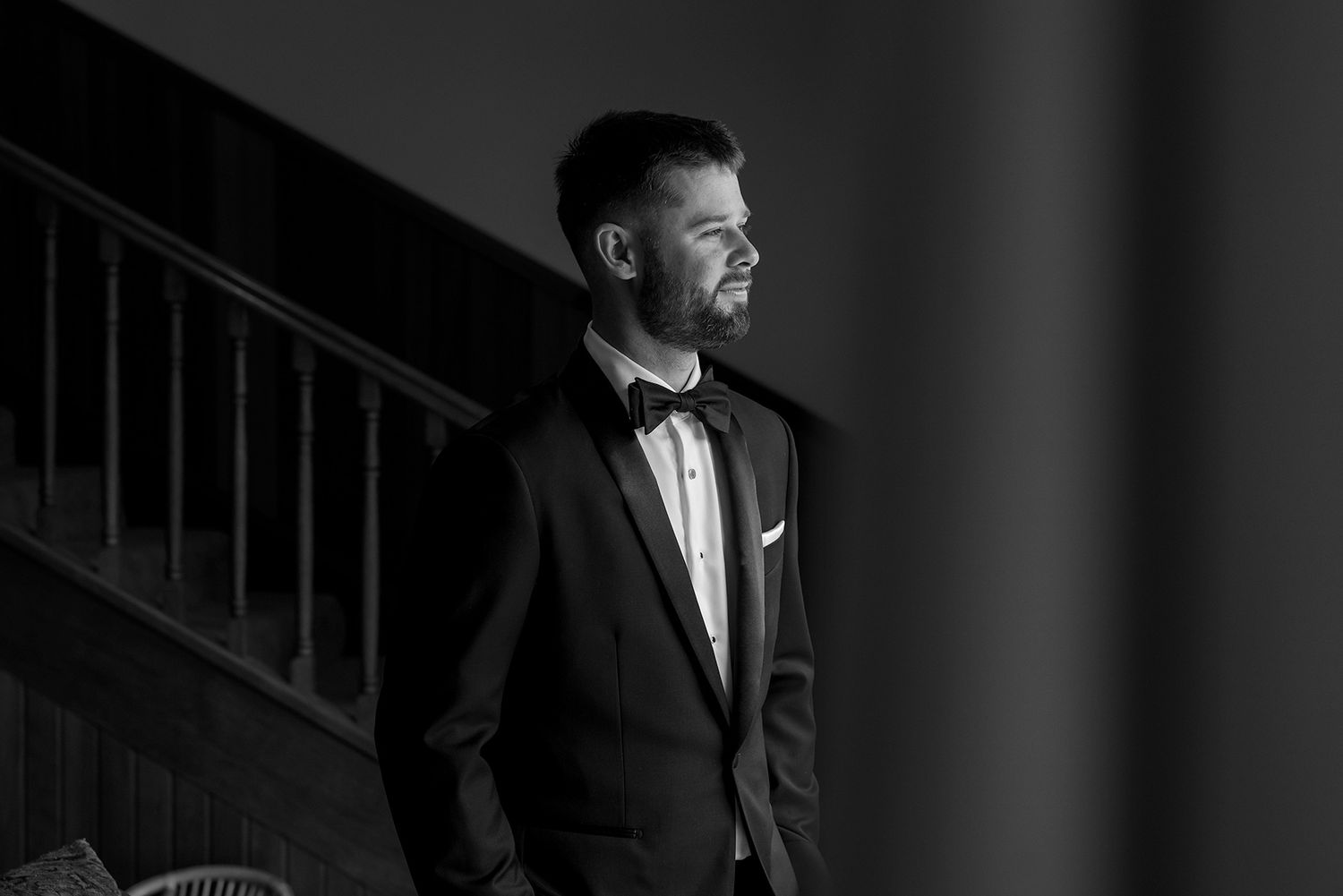 Black and white portrait of a Groom in a tuxedo bow tie and suit standing by a staircase before his wedding at Brown Brothers Winery.