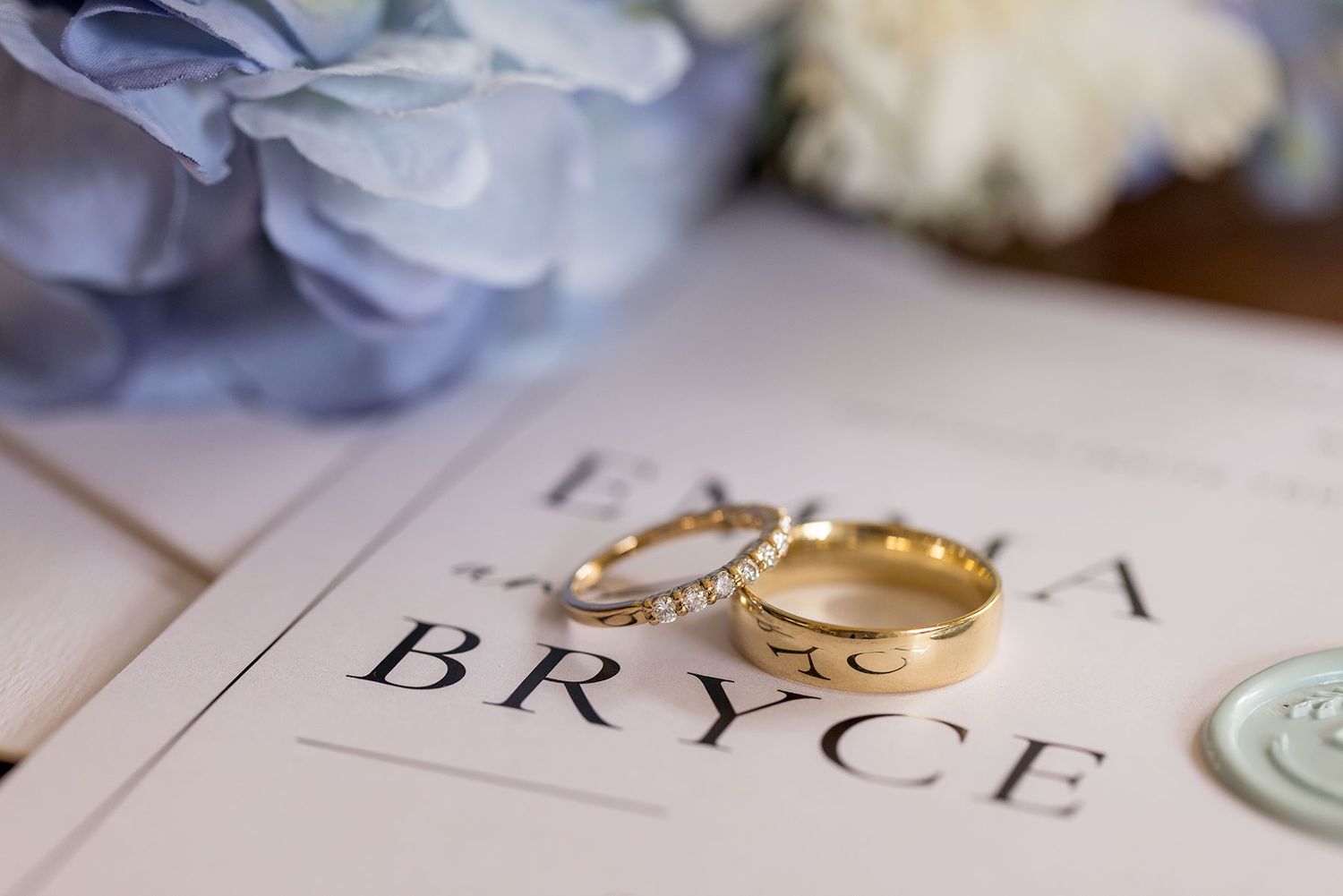 Two gold wedding rings rest on a paper or book page displaying the word BRYCE near blue floral decor.
