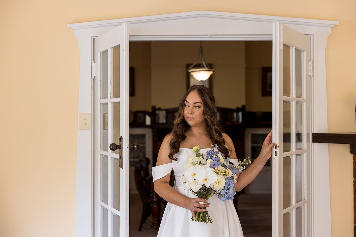 A bride in an off-shoulder white dress holds a bouquet while standing in a French door frame with natural light at Brown Brothers Winery.