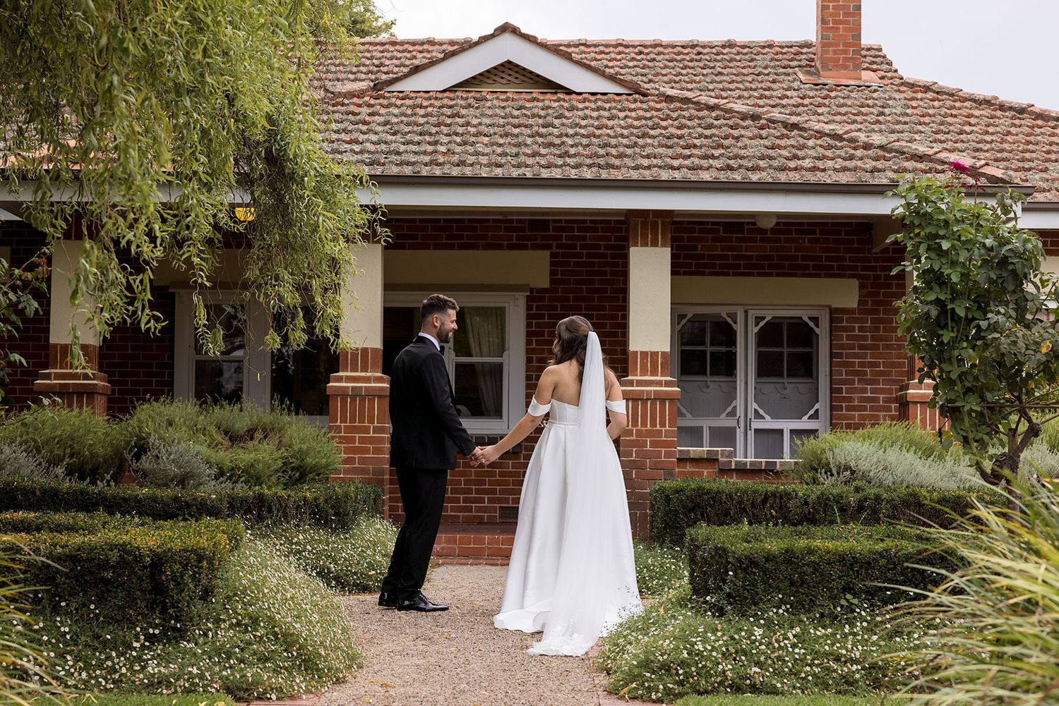 A couple shares their first look on their wedding day in front of a charming brick bungalow with a wraparound porch at Brown Brothers Winery