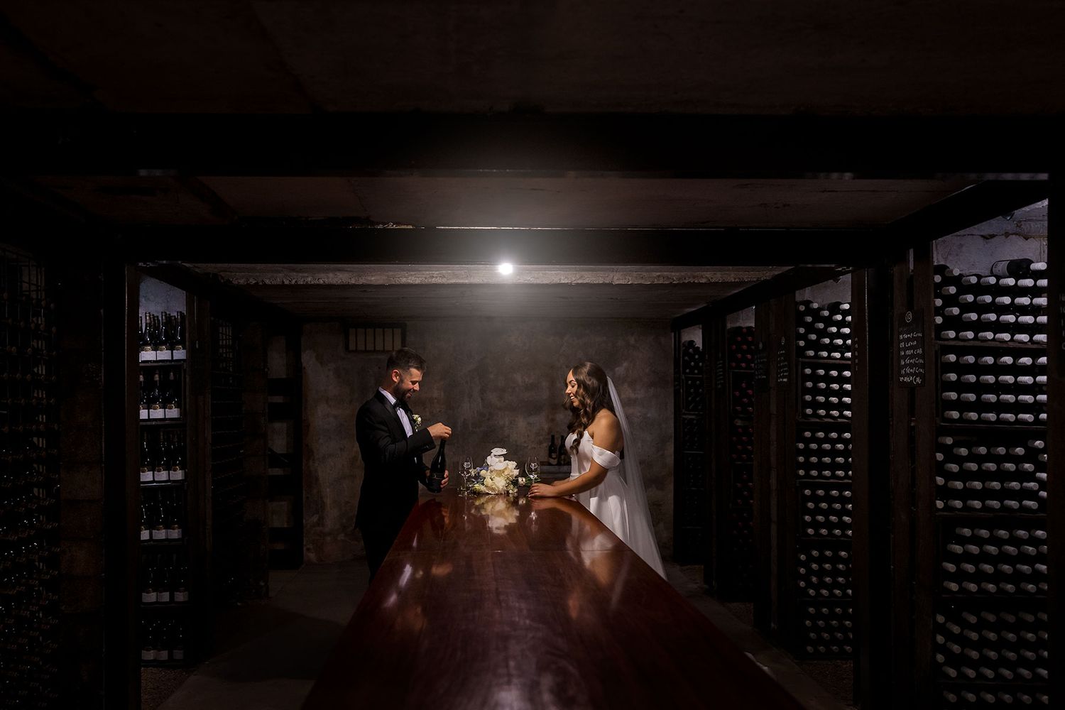 A couple stands together in a romantic, dimly lit wine cellar with rows of bottles creating a dramatic ambiance at Brown Brothers Winery.