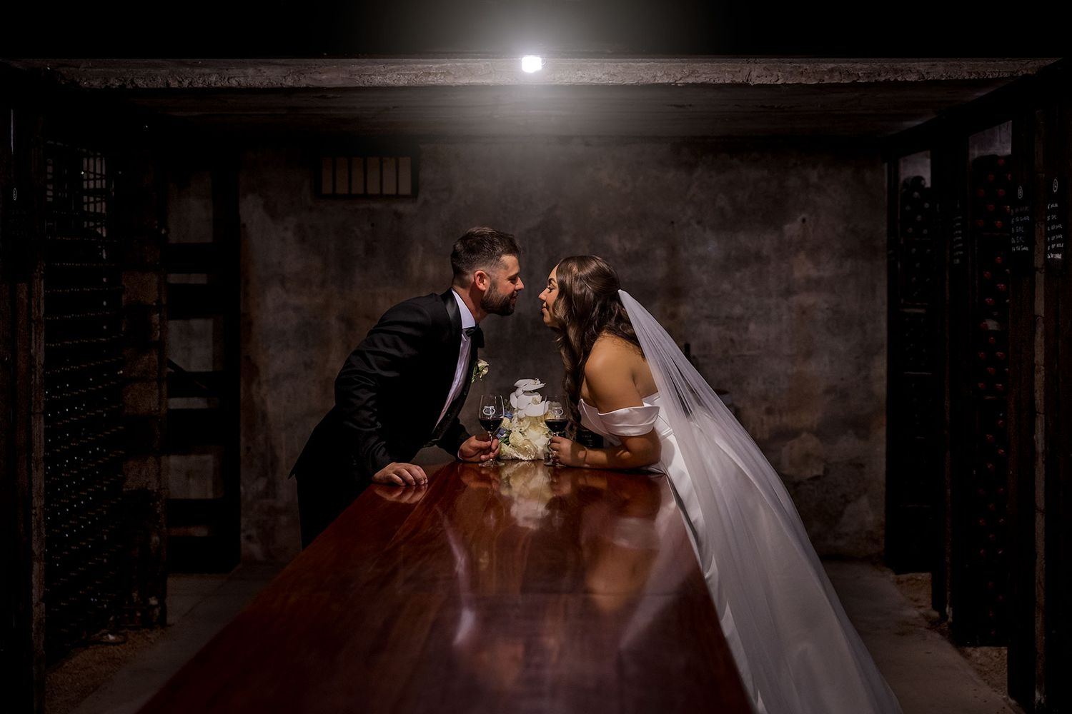 A couple stands together in a romantic, dimly lit wine cellar with rows of bottles creating a dramatic ambiance at Brown Brothers Winery.