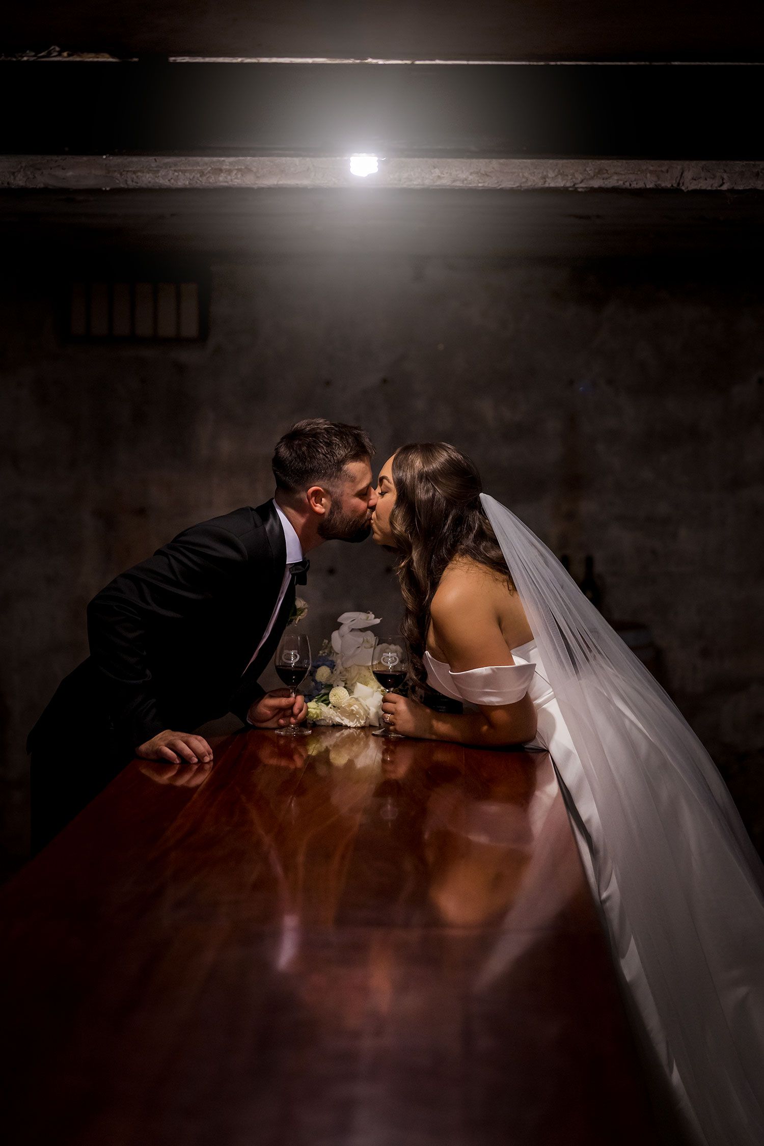 A couple stands together in a romantic, dimly lit wine cellar with rows of bottles creating a dramatic ambiance at Brown Brothers Winery.