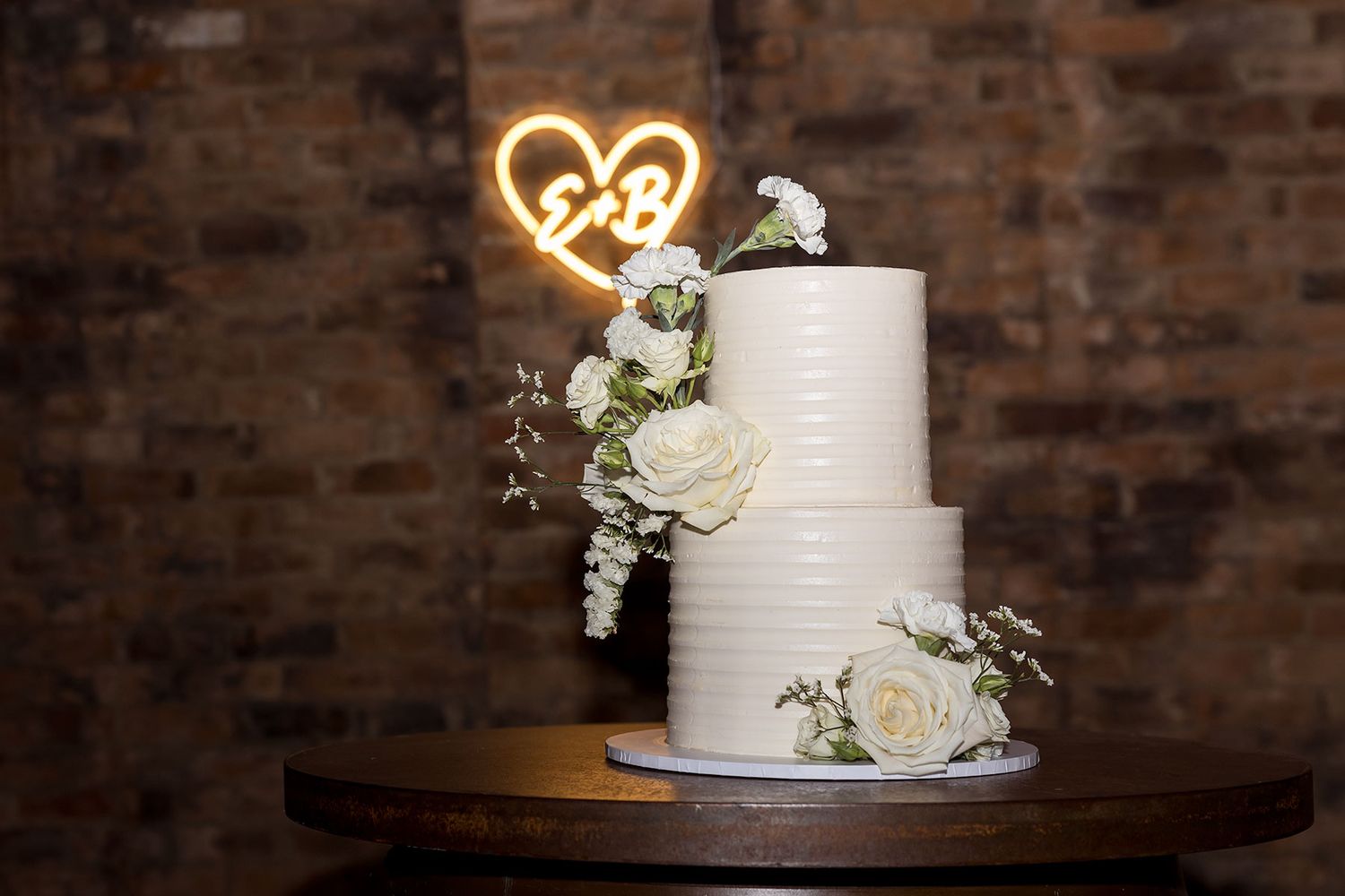 White wedding cake with a neon heart light displayed against rustic brick wall backdrop in the historic barn at Brown Brothers Winery.