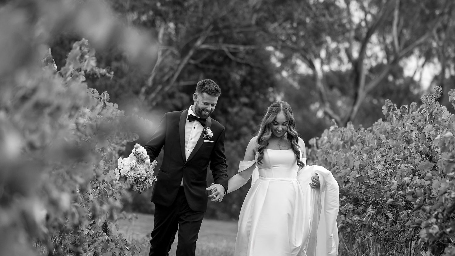 Romantic couple in wedding attire walk together holding hands in the vines at Brown Brothers Winery captured in Black and White.